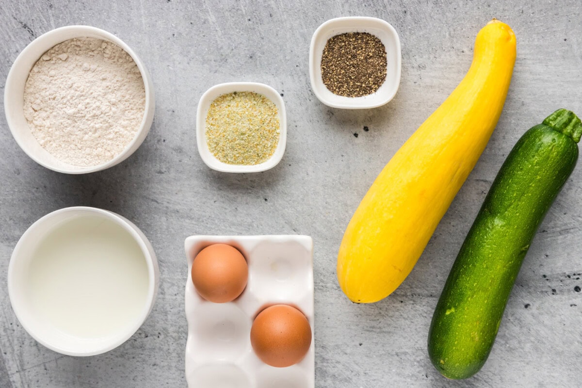 A kitchen counter with yellow squash, cornmeal, flour, eggs, buttermilk, kosher salt, black pepper, and garlic powder laid out for breading