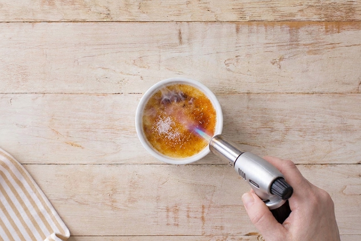 A kitchen torch caramelizing sugar on top of a chilled crème brûlée in a white ramekin