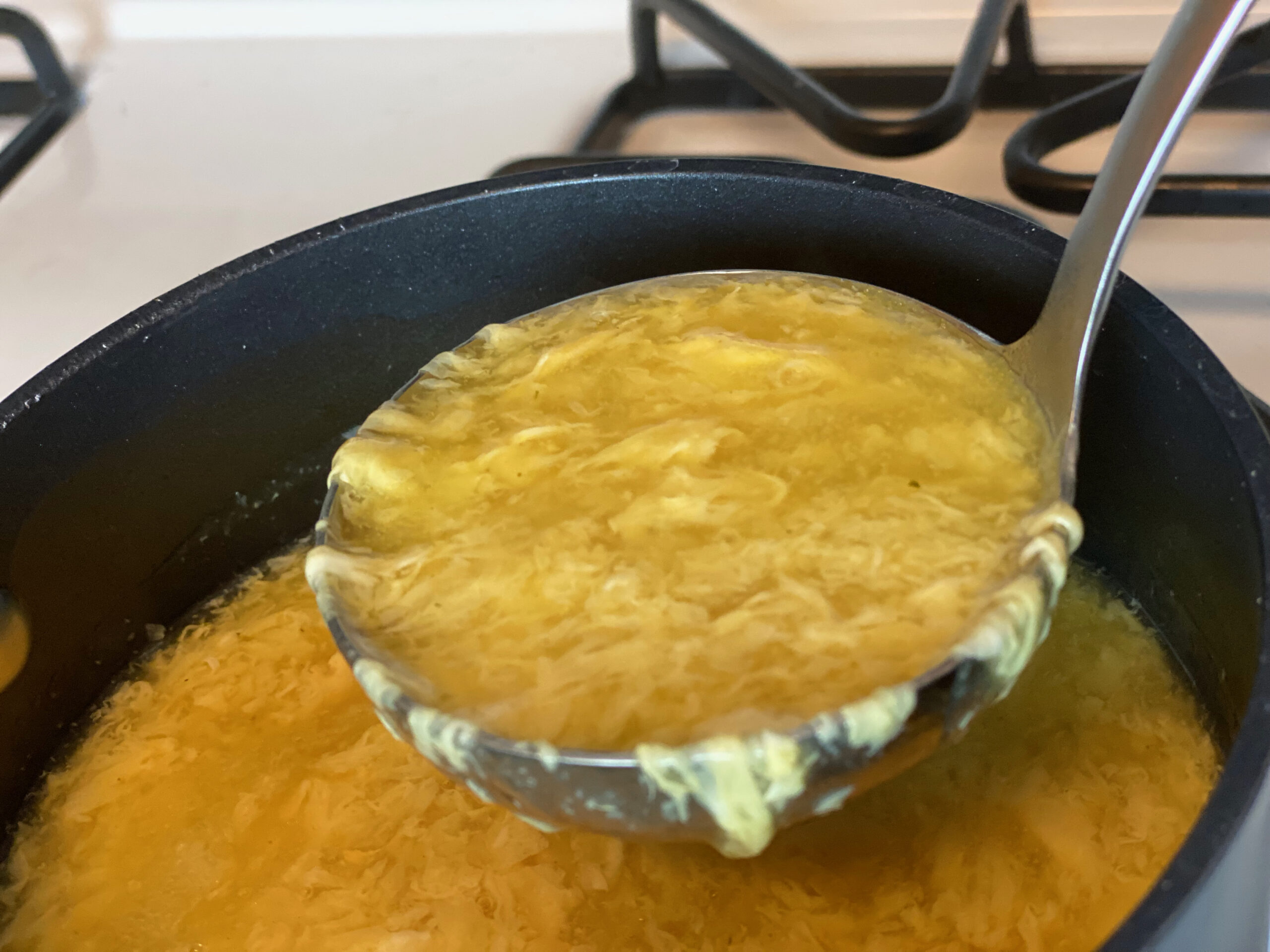 A ladle lifting egg drop soup from a small pot, showing delicate egg ribbons and a glossy golden broth