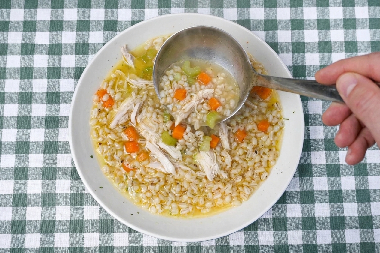 A ladle pouring chicken barley soup into a bowl with visible carrots, celery, shredded chicken, and pearled barley