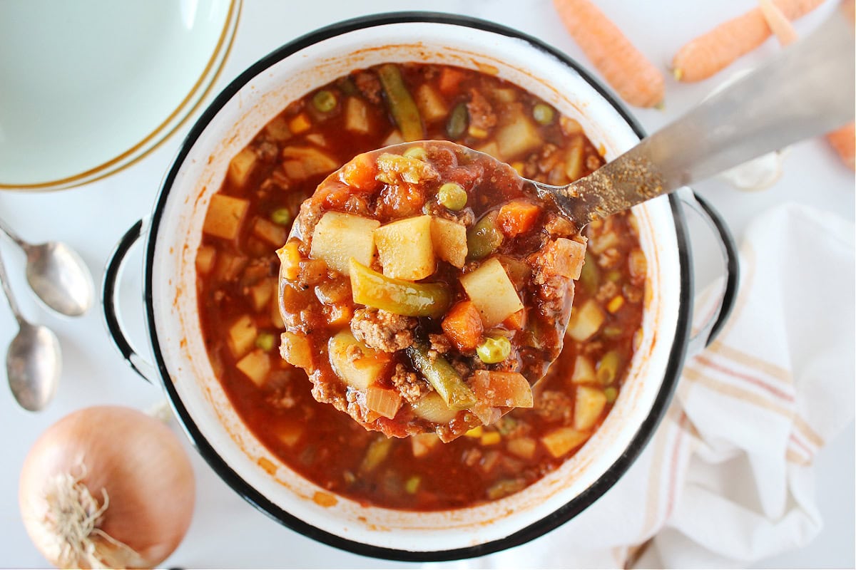 A ladle pouring hot hamburger stew into a ceramic bowl on a wooden table with a spoon beside it