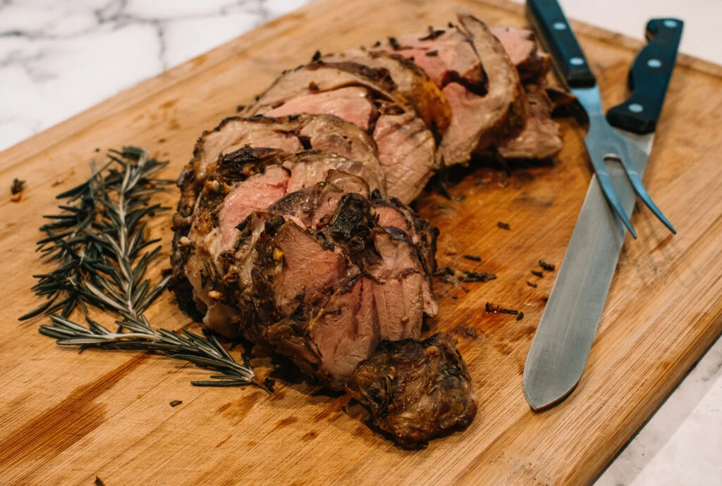 A lamb thigh roast resting on a wooden cutting board with foil loosely tented, with a small saucepan of pan sauce nearby on the same kitchen counter