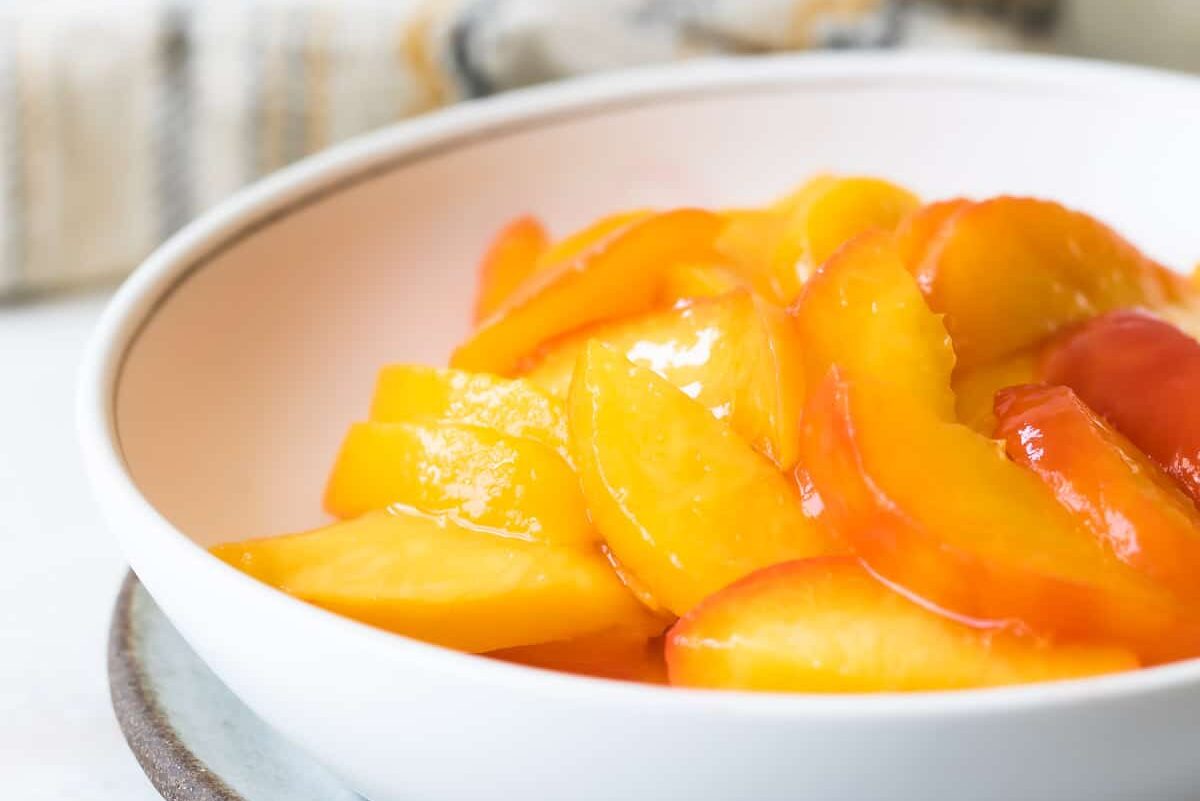 A large bowl filled with peeled, freshly sliced peaches on a kitchen counter beside measuring spoons and a lemon