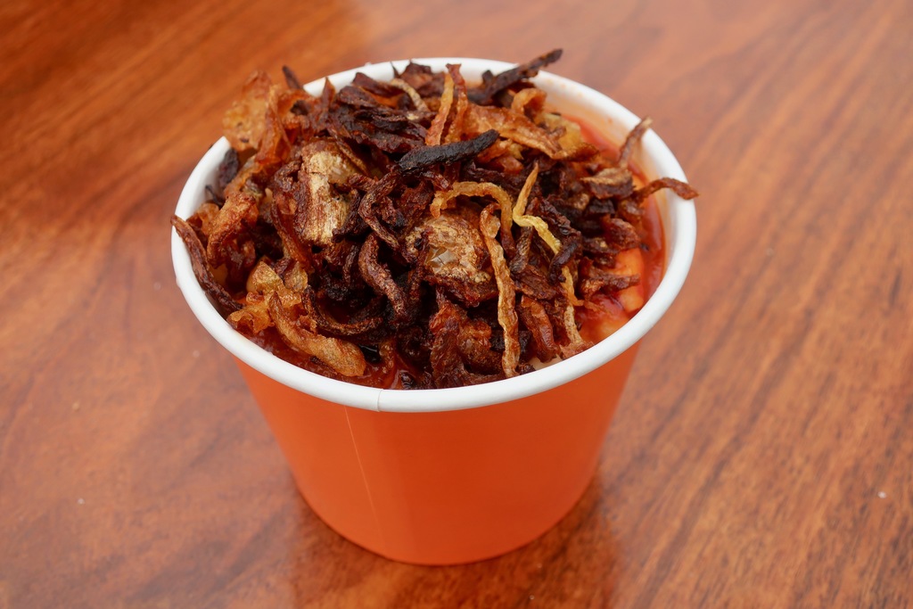 A large bowl of Egyptian koshari with layers of rice, lentils, and pasta topped with crispy fried onions and spooned-over tomato sauce on a warm-toned kitchen table