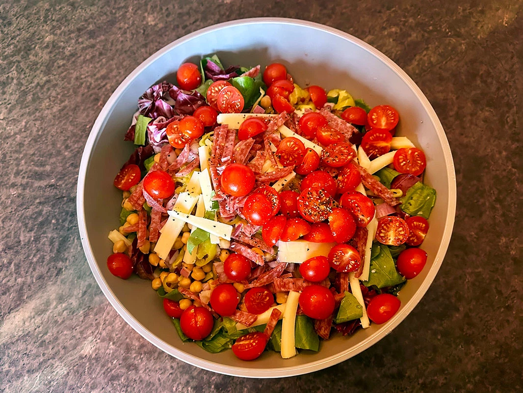 A large bowl of Italian chopped salad with romaine, salami, provolone cubes, chickpeas, cherry tomatoes, red onion, pepperoncini, and herbs on a bright kitchen counter