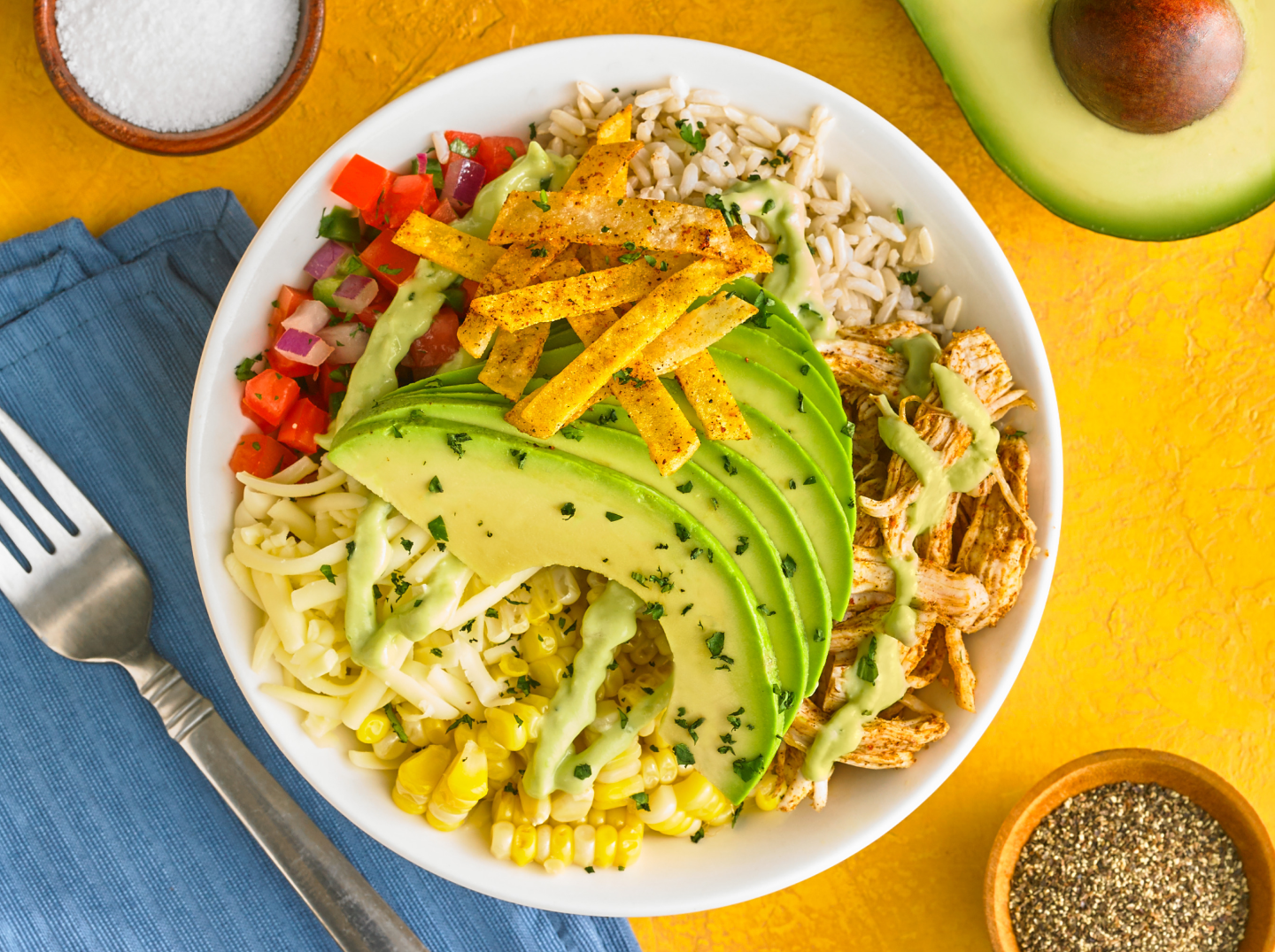 A large bowl of Southwest salad with romaine, black beans, corn, avocado slices, cherry tomatoes, red onion, tortilla strips, and a creamy lime dressing drizzled over the top on a bright kitchen table