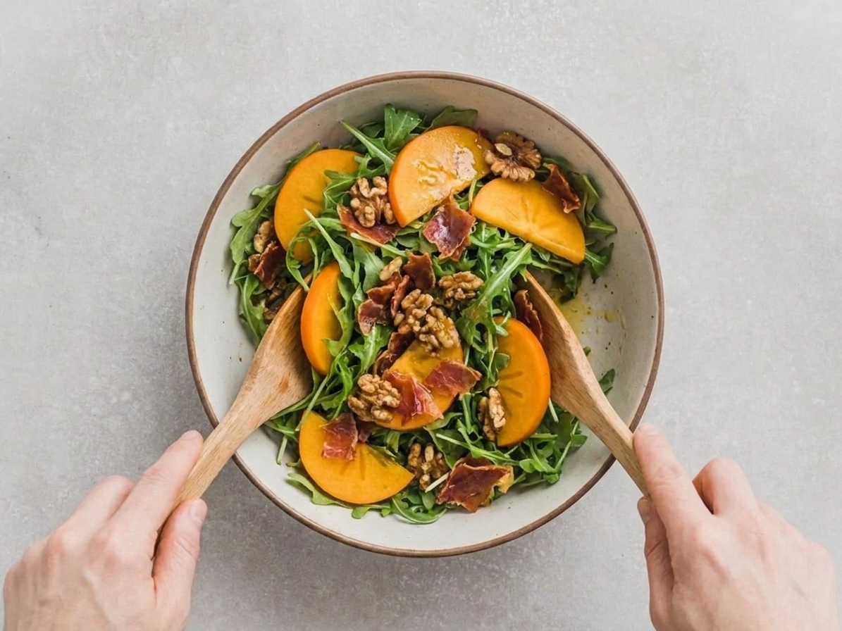 A large bowl of arugula and persimmon being gently tossed with vinaigrette using salad tongs