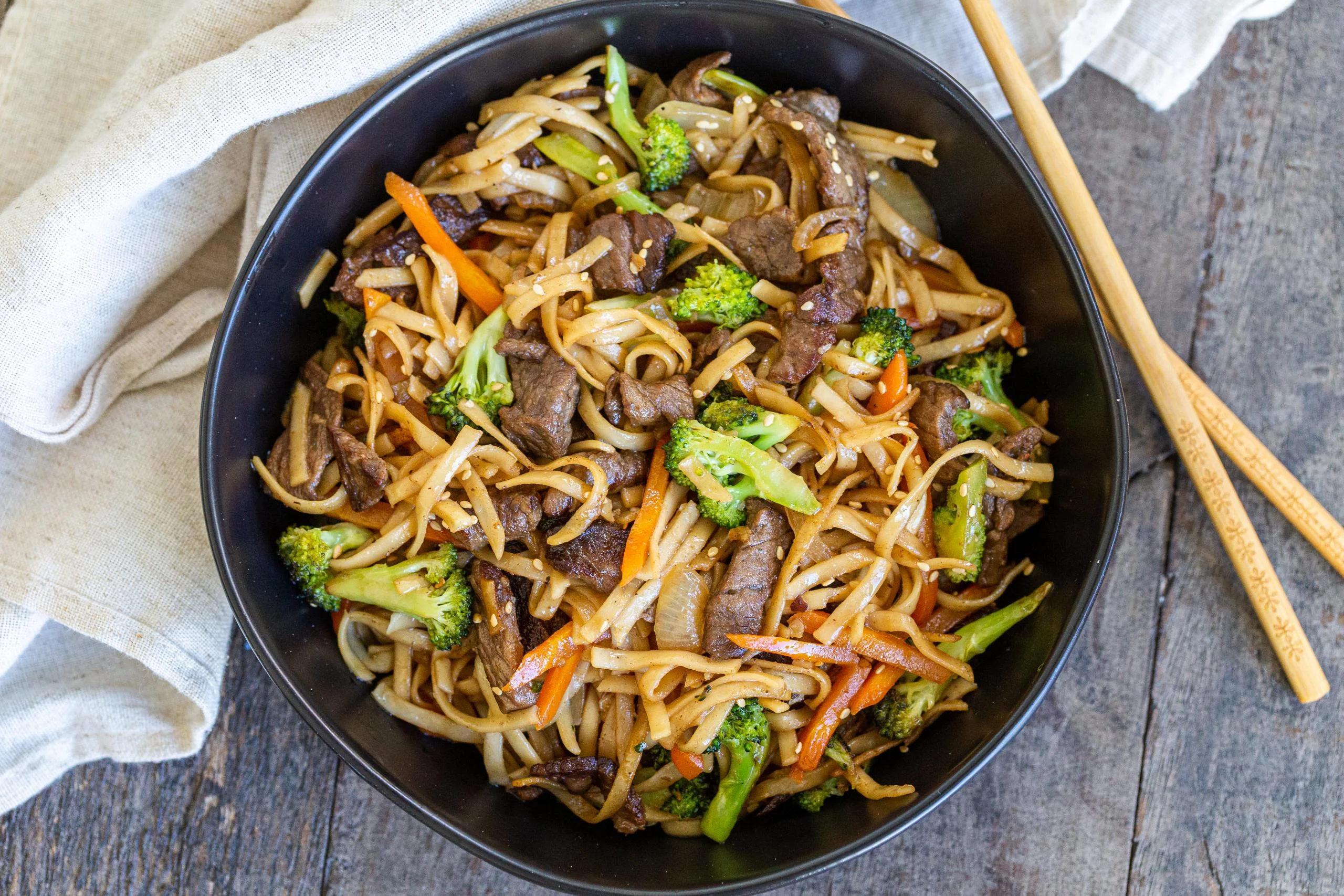 A large bowl of beef lo mein with glossy noodles, seared slices of beef, broccoli, carrots, and scallions on a wooden table with chopsticks