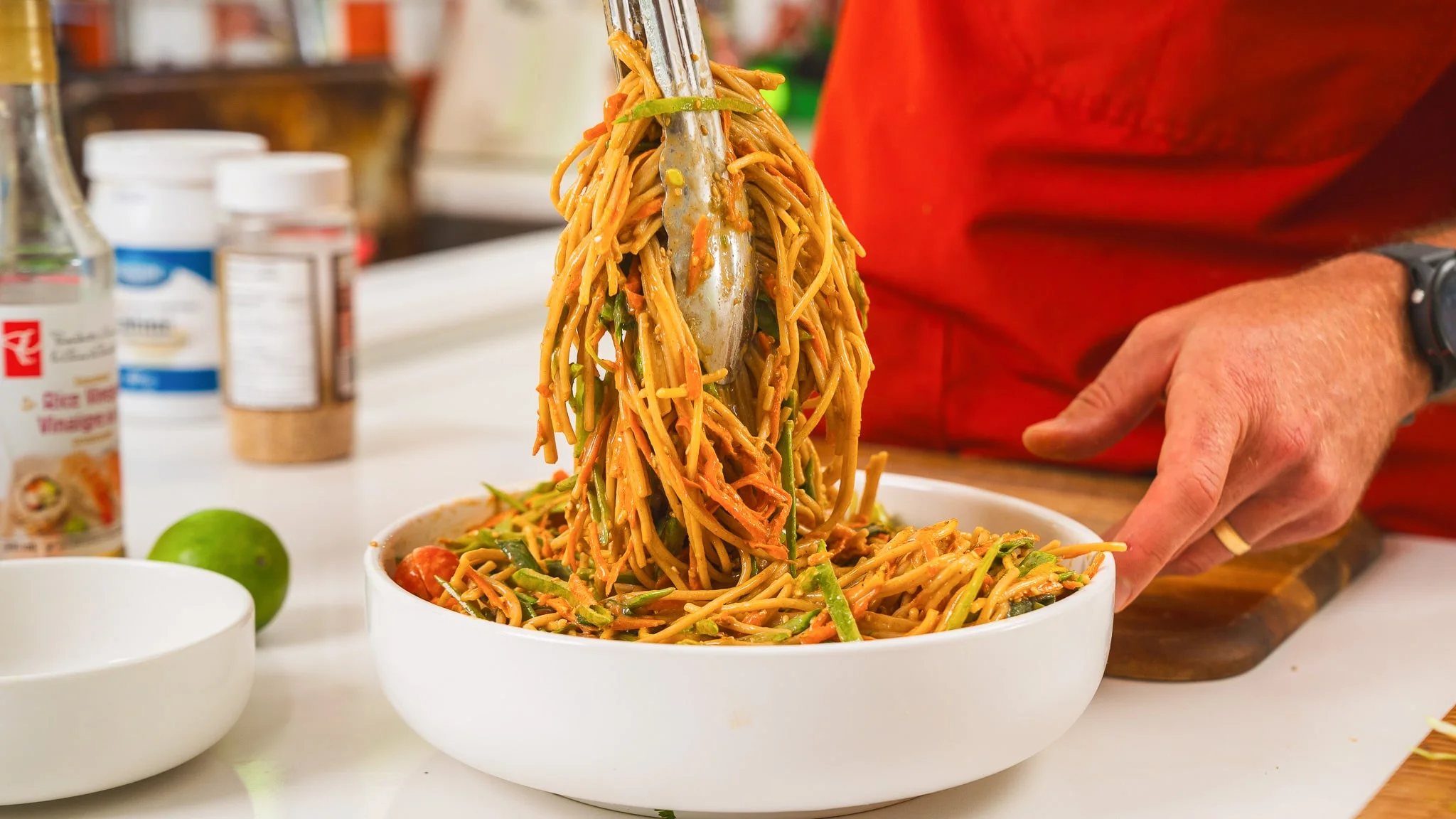 A large bowl of cooked noodles being tossed with sesame sauce using tongs on a kitchen counter