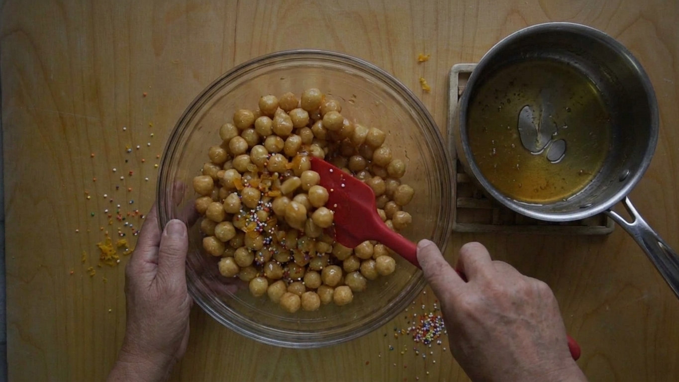 A large bowl of fried struffoli being tossed with warm honey glaze using a silicone spatula on a kitchen counter