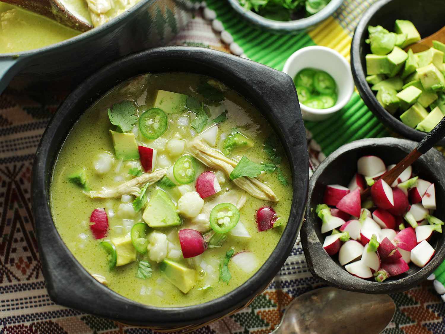 A large bowl of pozole verde with chicken and hominy, topped with shredded cabbage, sliced radishes, cilantro, and a lime wedge on a warm kitchen table