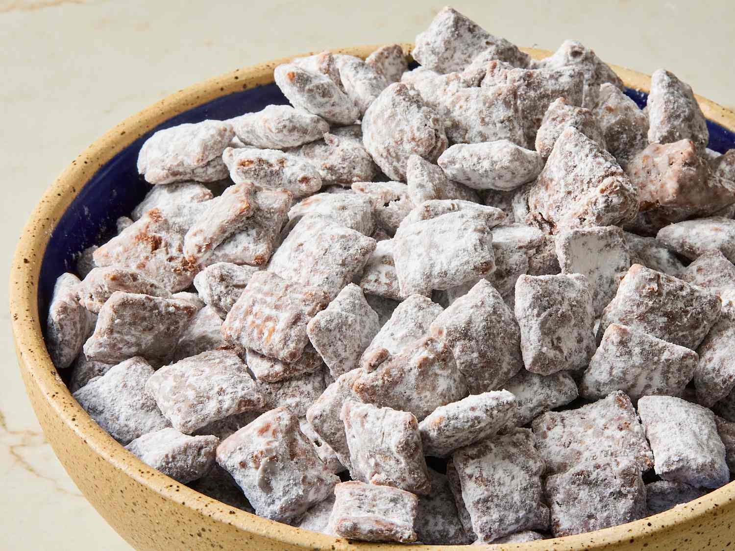 A large bowl of puppy chow made with Chex cereal, heavily dusted with powdered sugar, sitting on a warm kitchen counter with soft natural light