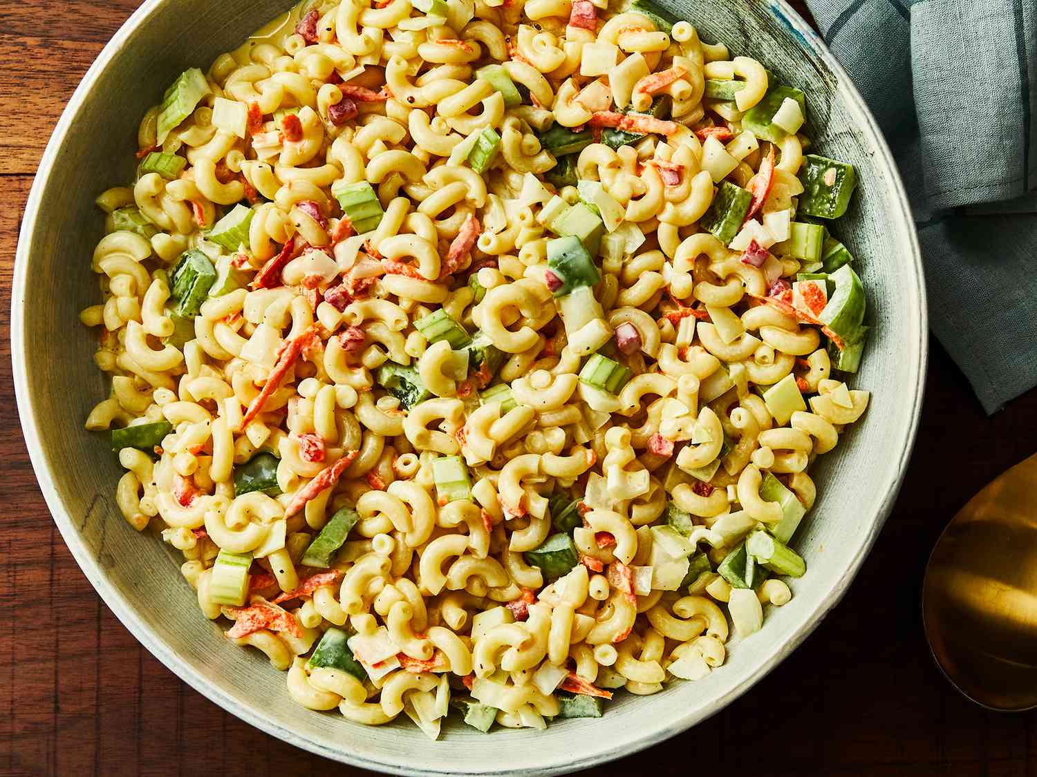 A large ceramic bowl filled with creamy classic macaroni salad with celery, red onion, shredded carrot, and small pickle pieces, set on a bright kitchen counter with a serving spoon