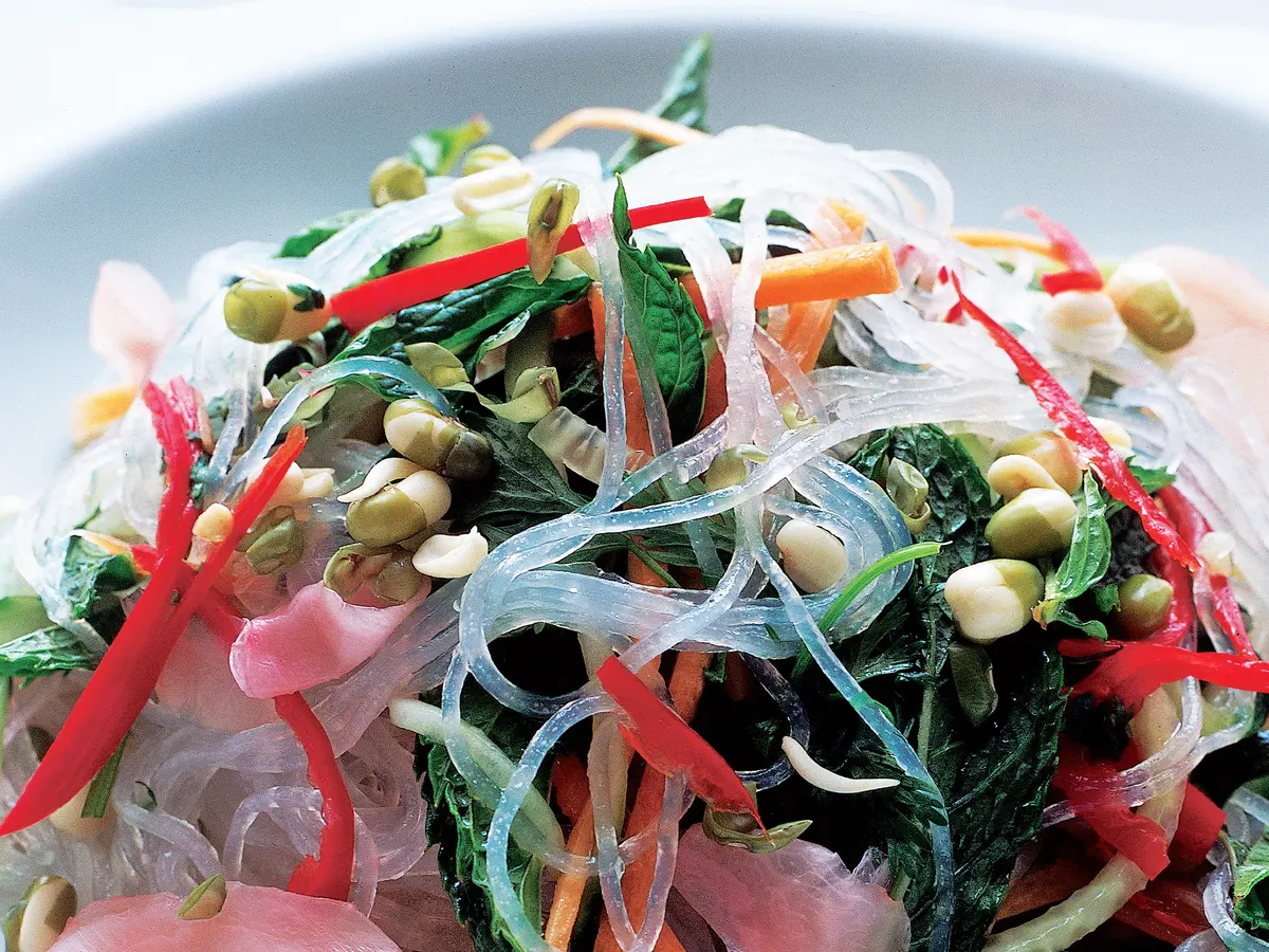A large ceramic bowl filled with crystal noodles tossed with cucumbers, carrots, herbs, and sesame, with a small bowl of lime soy dressing nearby on a light kitchen table