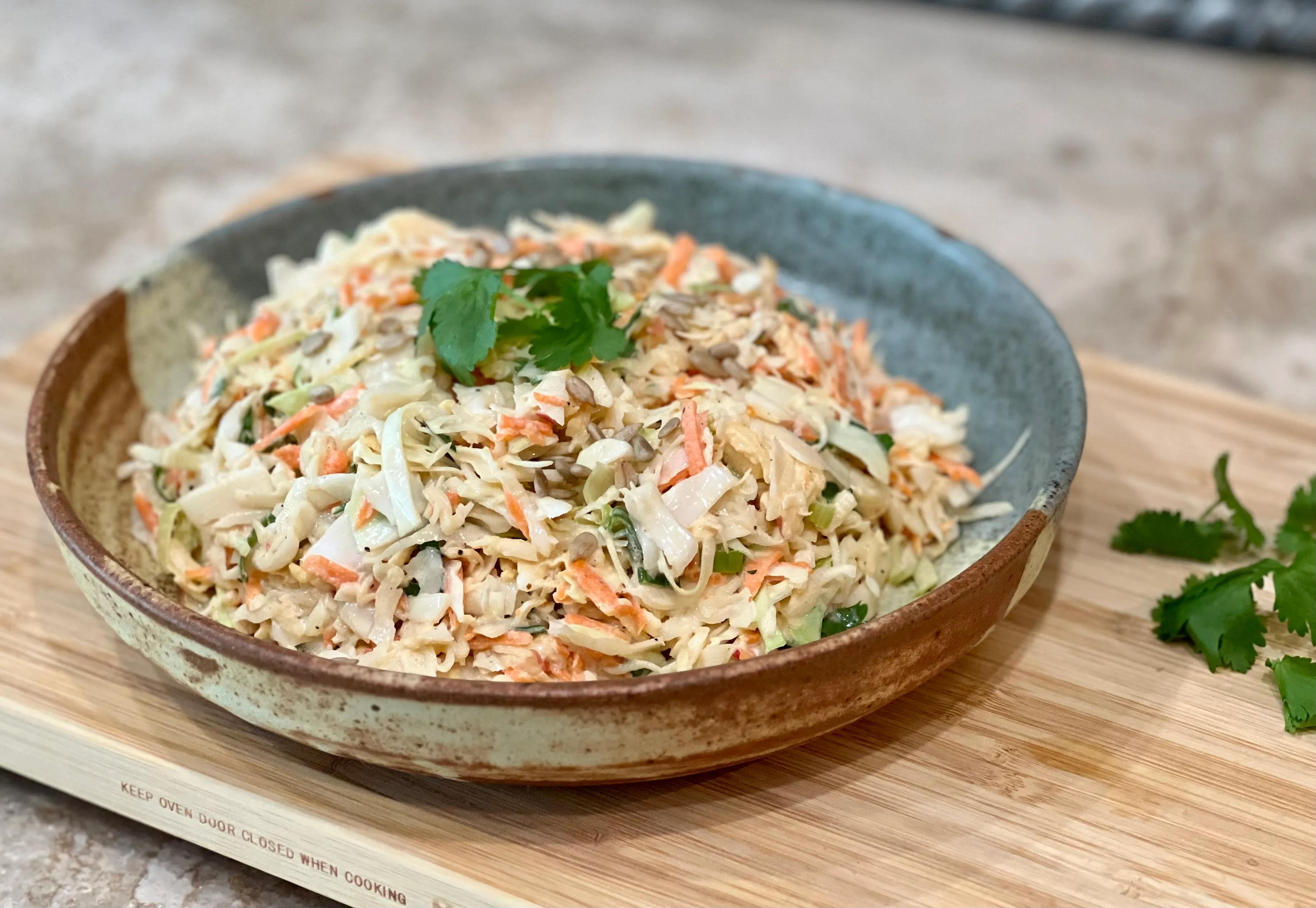 A large ceramic bowl of creamy coleslaw with shredded green cabbage, carrots, and black pepper on a wooden kitchen table with soft natural light