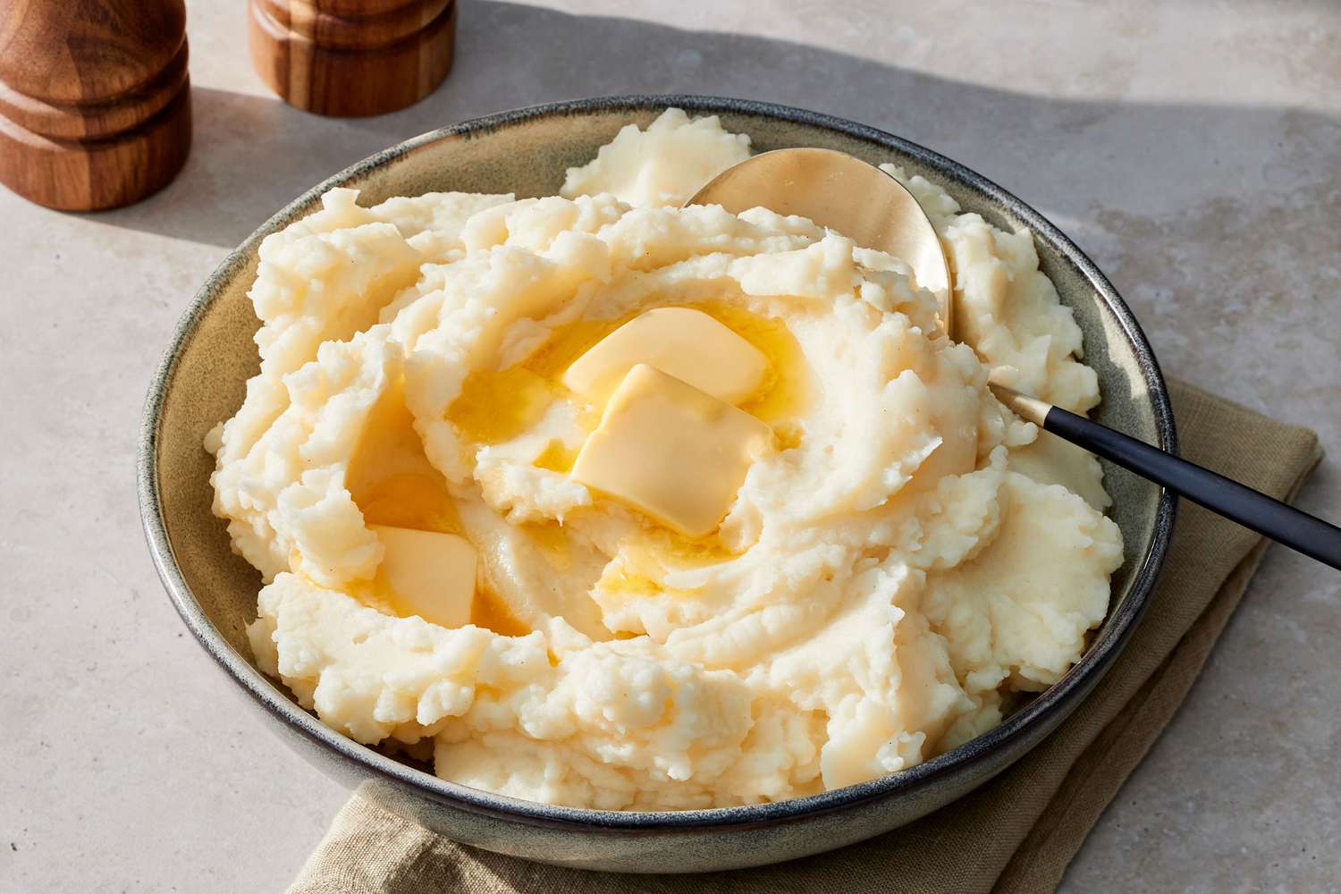 A large ceramic bowl of creamy mashed potatoes with a swirl of melted butter on top, flaky salt, and chopped chives on a wooden table with soft natural kitchen light