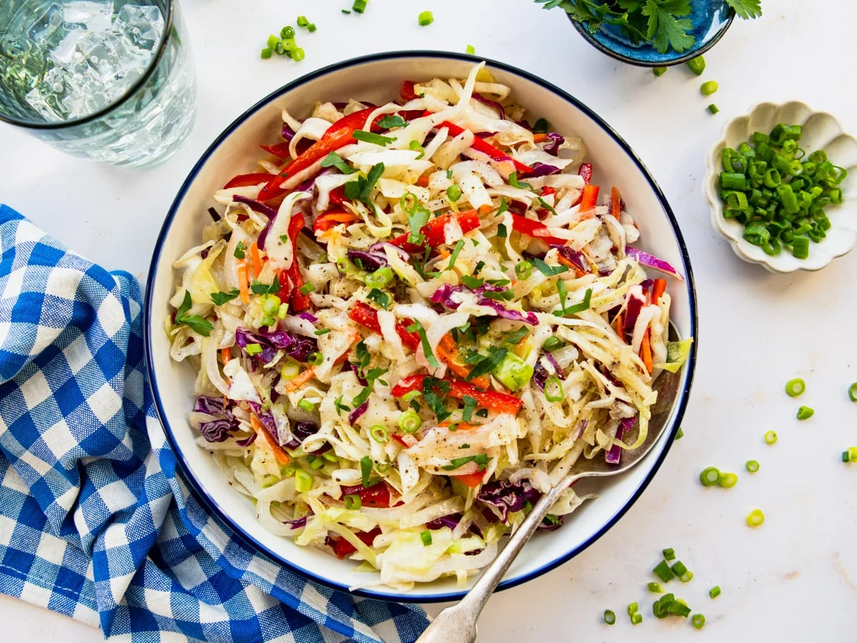 A large ceramic bowl of vinegar-based coleslaw with shredded green cabbage, purple cabbage, and carrots, with a light glossy dressing on a bright kitchen counter in soft natural light