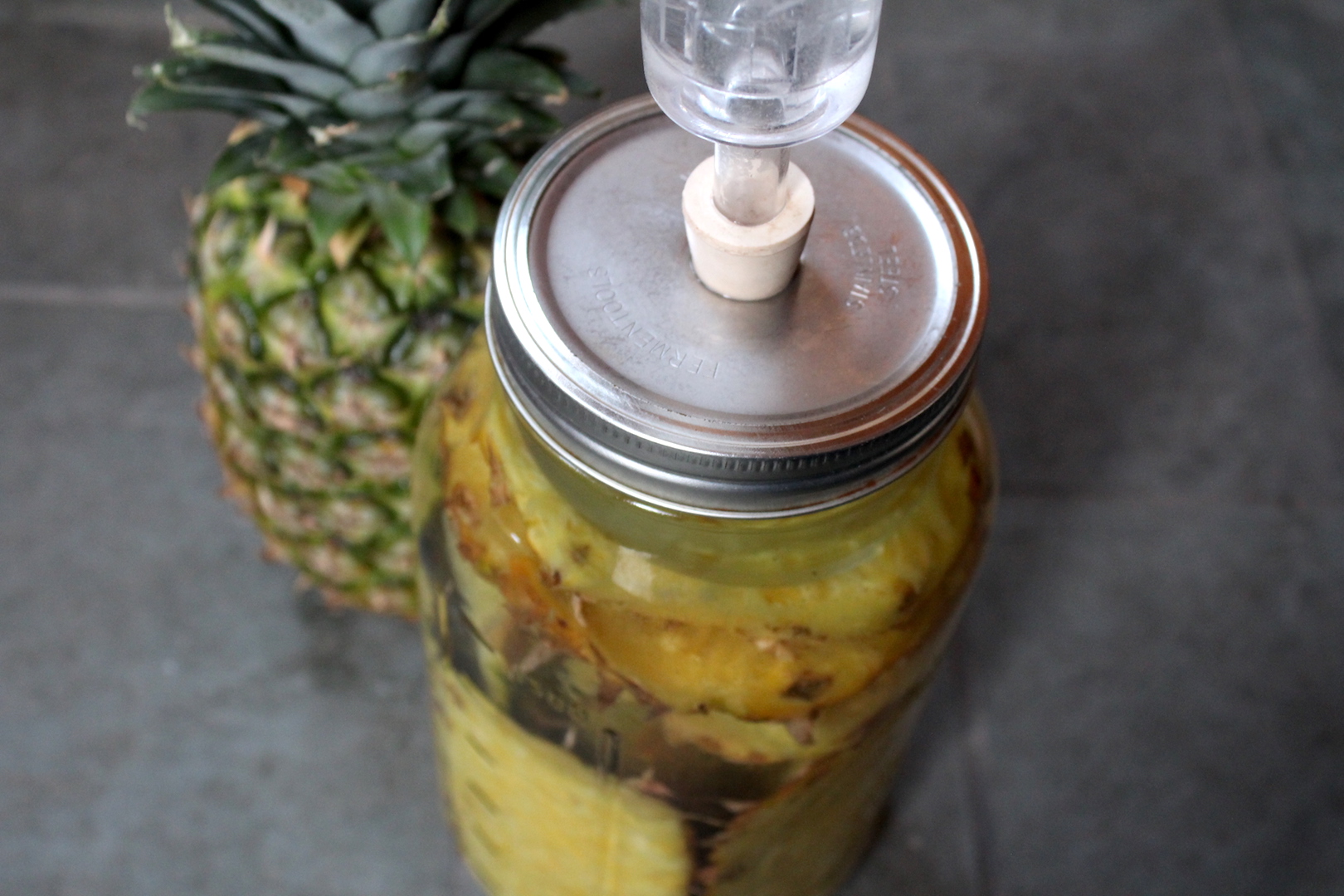 A large glass jar of tepache on a kitchen counter with pineapple peels floating on top and a small bowl of piloncillo nearby, warm natural light