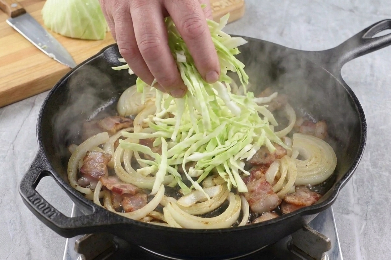 A large handful of sliced cabbage being added to a hot skillet with bacon and onions, steam rising
