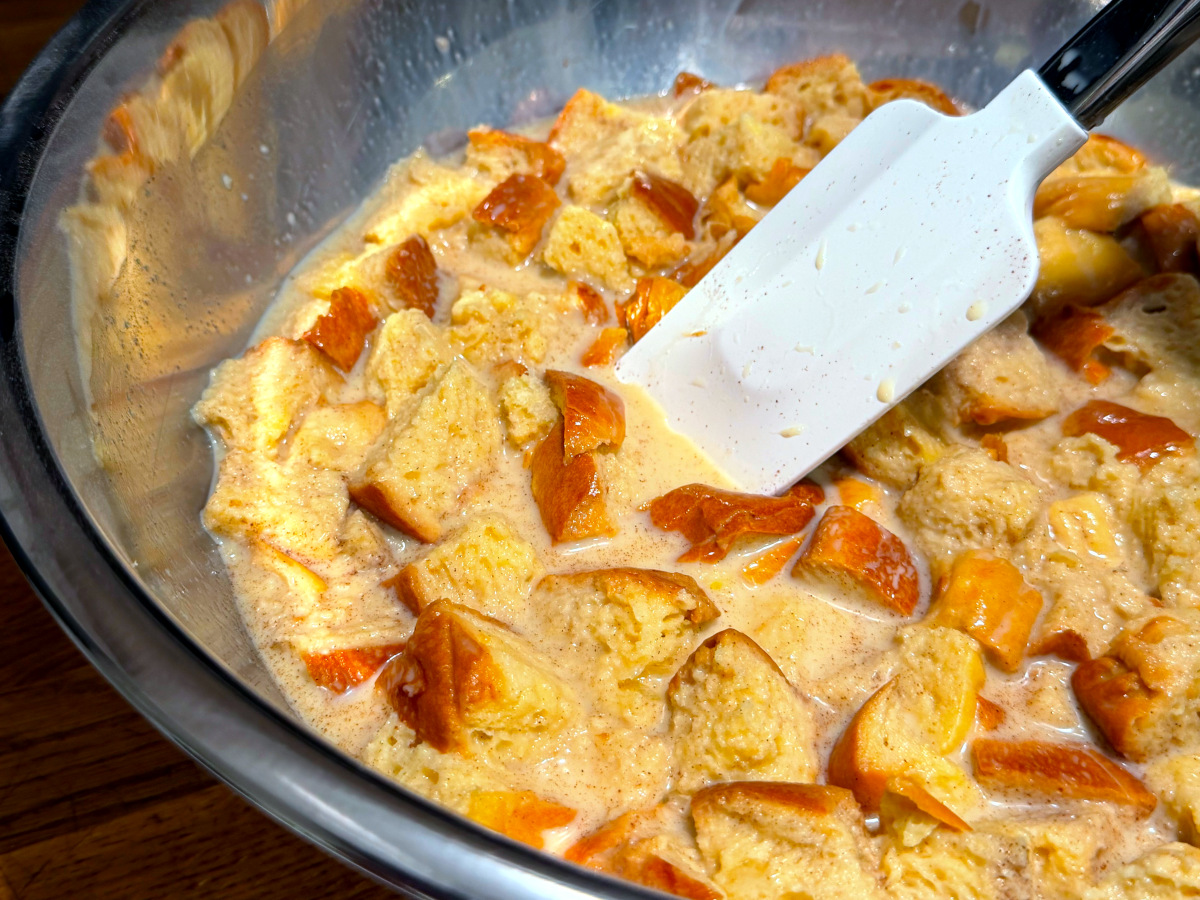 A large mixing bowl filled with torn bread cubes soaking in a pale custard with cinnamon specks, a wooden spoon resting on the rim, real kitchen photo