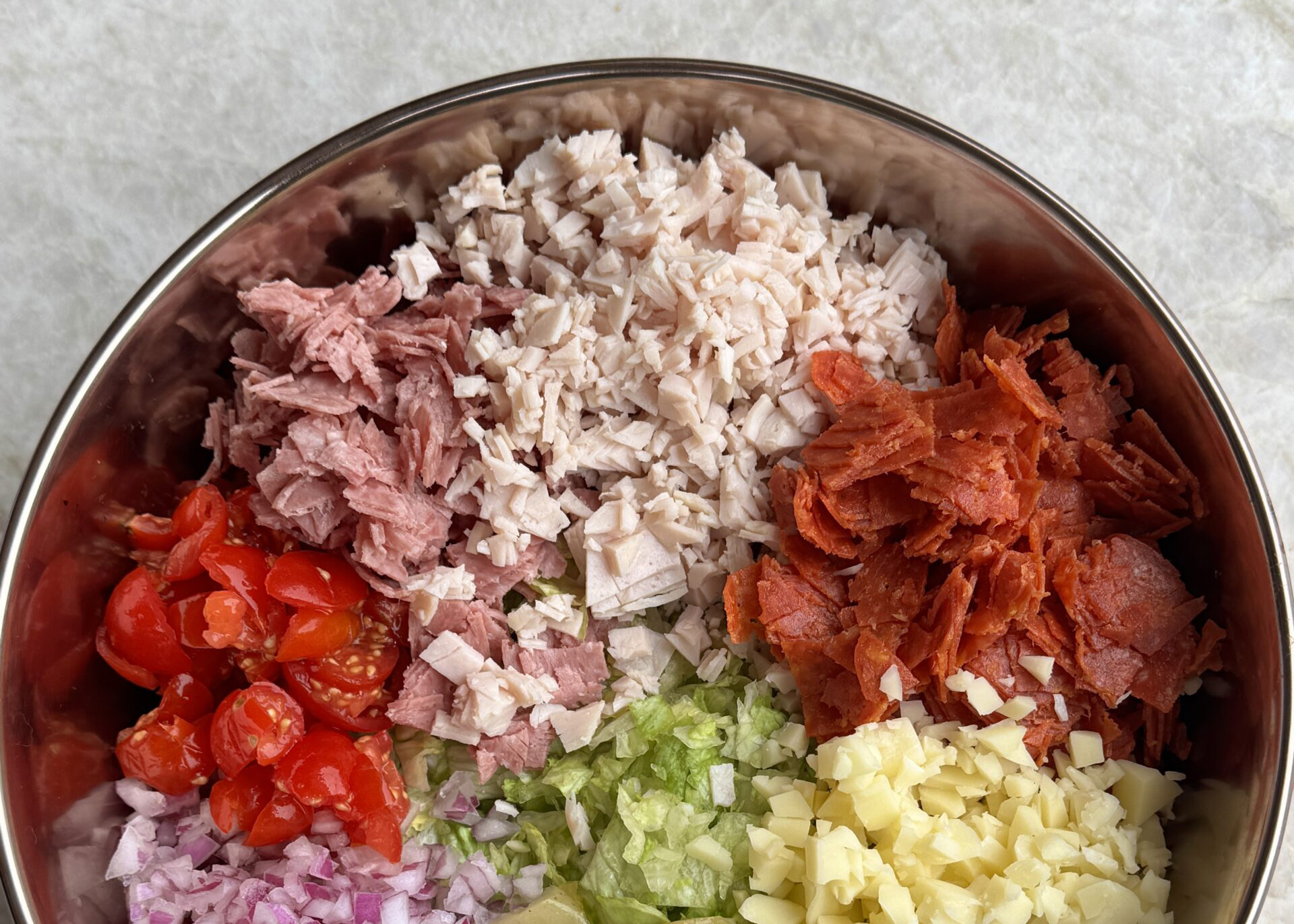 A large mixing bowl on a kitchen counter with shredded iceberg lettuce, deli meats, provolone, tomatoes, and pepperoncini being tossed together with dressing