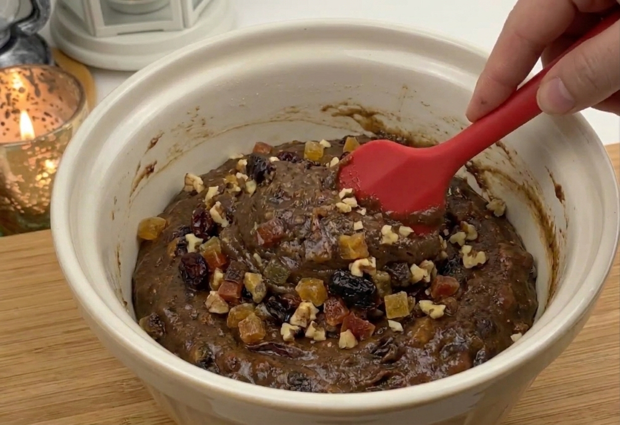 A large mixing bowl with thick fruit cake batter and a spatula, with visible dried fruit and chopped nuts on a kitchen counter