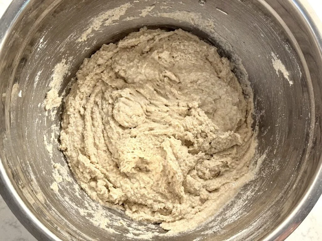 A large mixing bowl with thick gluten-free sourdough dough being stirred with a sturdy spatula on a kitchen counter