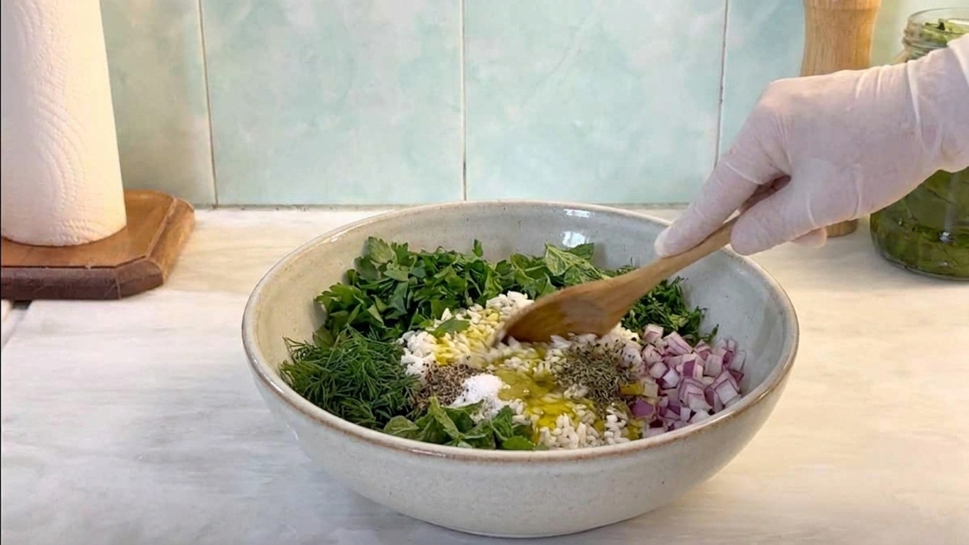 A large mixing bowl with uncooked rice, chopped herbs, diced onions, spices, olive oil, and lemon juice being stirred with a spoon on a kitchen counter