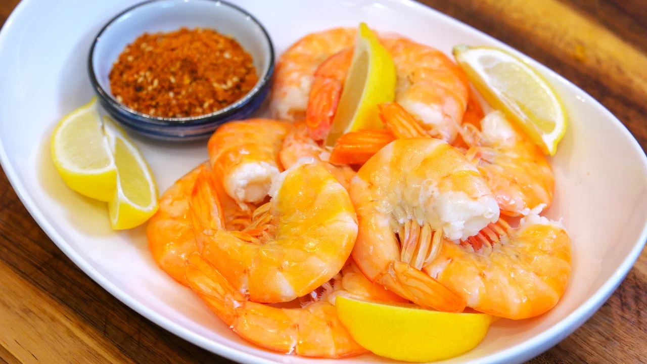 A large platter of chilled boiled shrimp arranged in a ring with lemon wedges and a small bowl of cocktail sauce on a wooden table