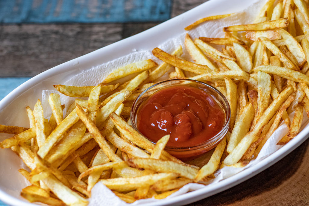 A large platter of golden homemade French fries with crisp edges and a small bowl of ketchup on a simple wooden table setting