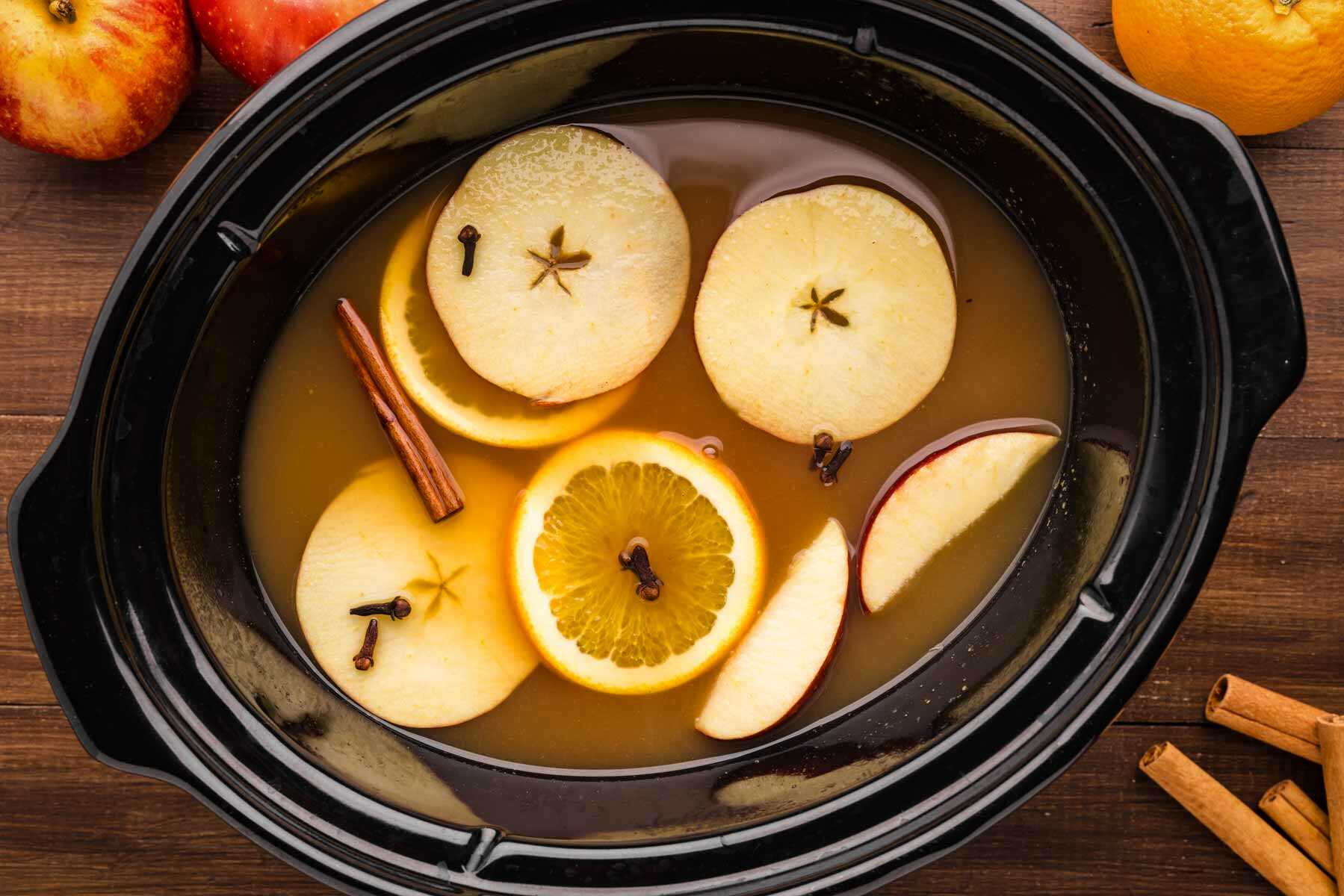 A large pot of hot apple cider simmering on a stovetop with cinnamon sticks and orange slices floating on top in a cozy home kitchen