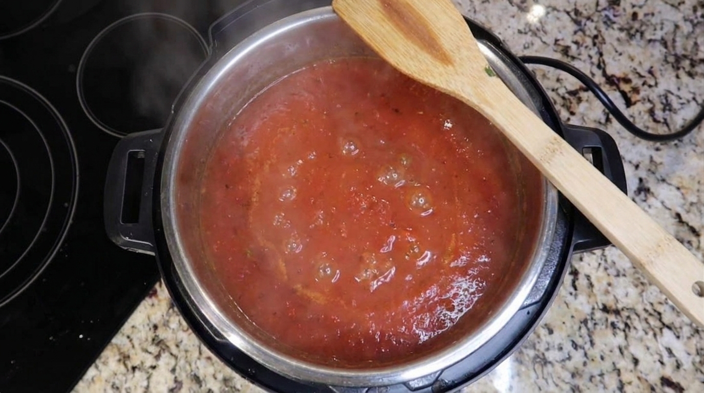 A large pot of tomato-based soup base simmering with small bubbles and a wooden spoon resting on the rim