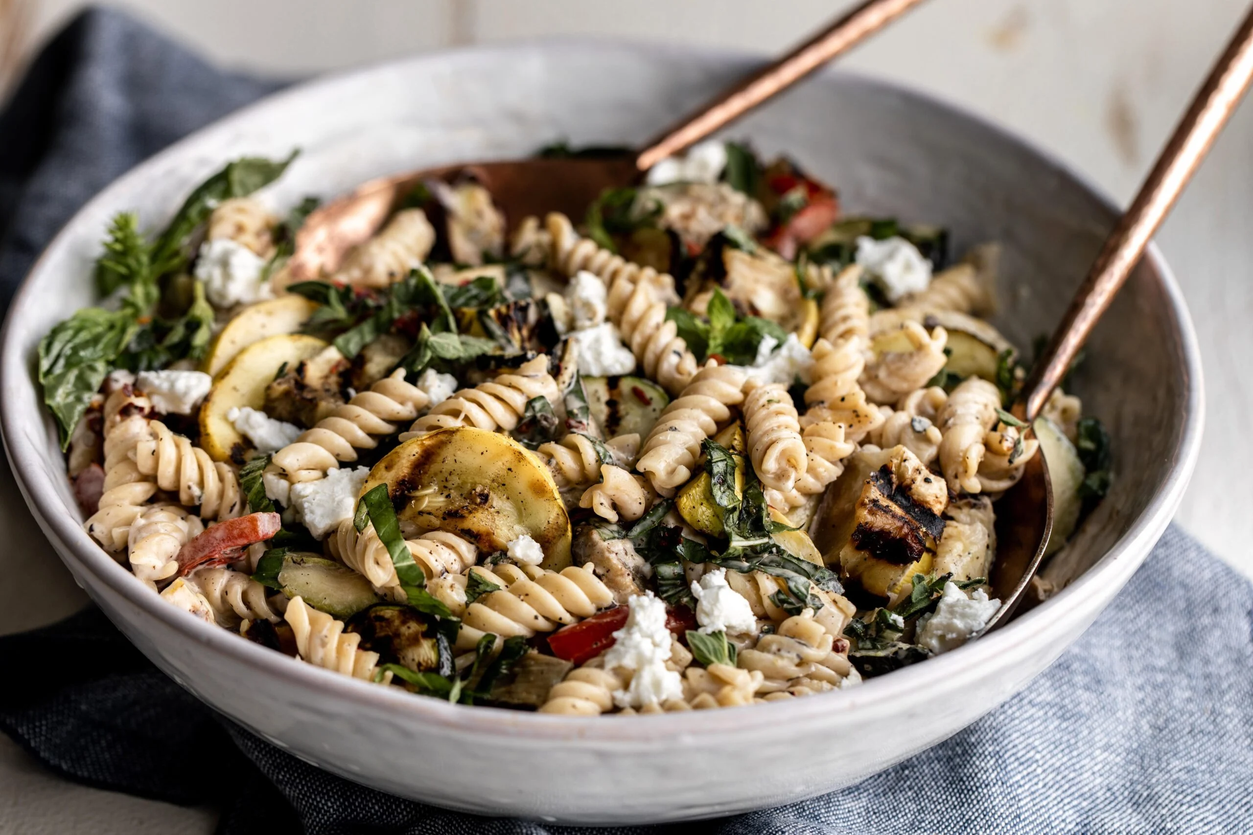 A large serving bowl filled with rotini pasta salad with cherry tomatoes, cucumber, bell pepper, cheddar cubes, and salami, tossed in a glossy Italian style dressing on a bright kitchen counter