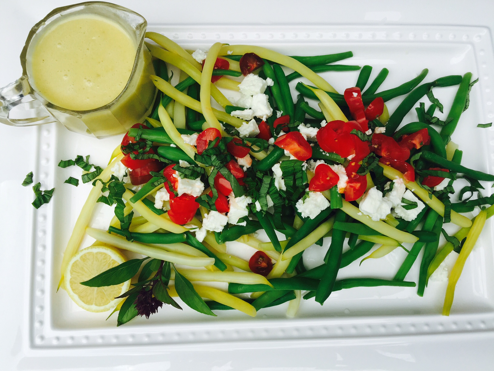 A large serving bowl of green bean salad with lemon slices, toasted almonds, shaved parmesan, and a glossy vinaigrette on a light kitchen counter