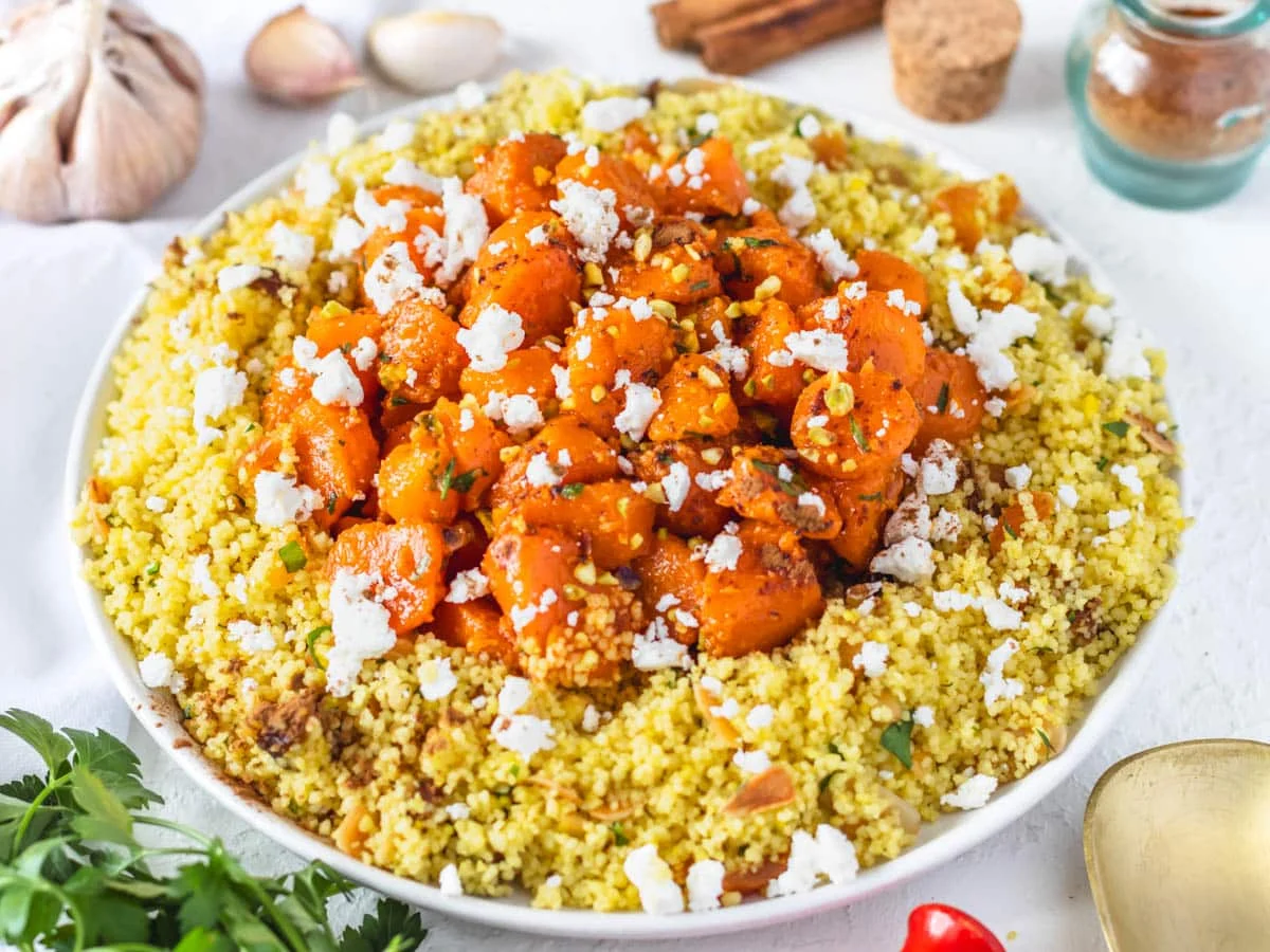 A large shallow bowl of Moroccan couscous with chickpeas, golden raisins, toasted almonds, and chopped herbs, set on a wooden table with warm natural light