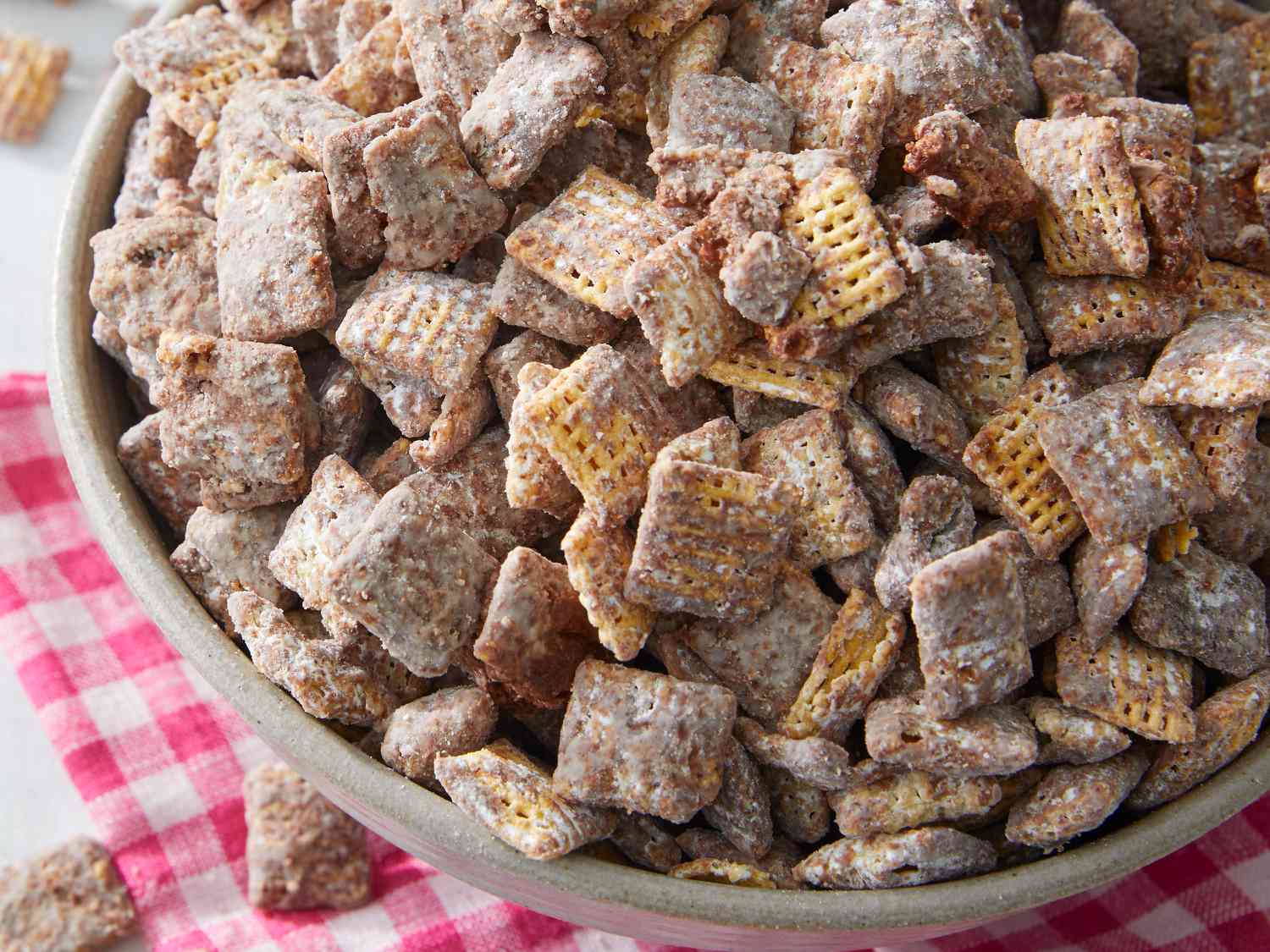 A large white bowl filled with muddy buddy snack mix, coated in powdered sugar, on a light kitchen counter with a wooden spoon nearby