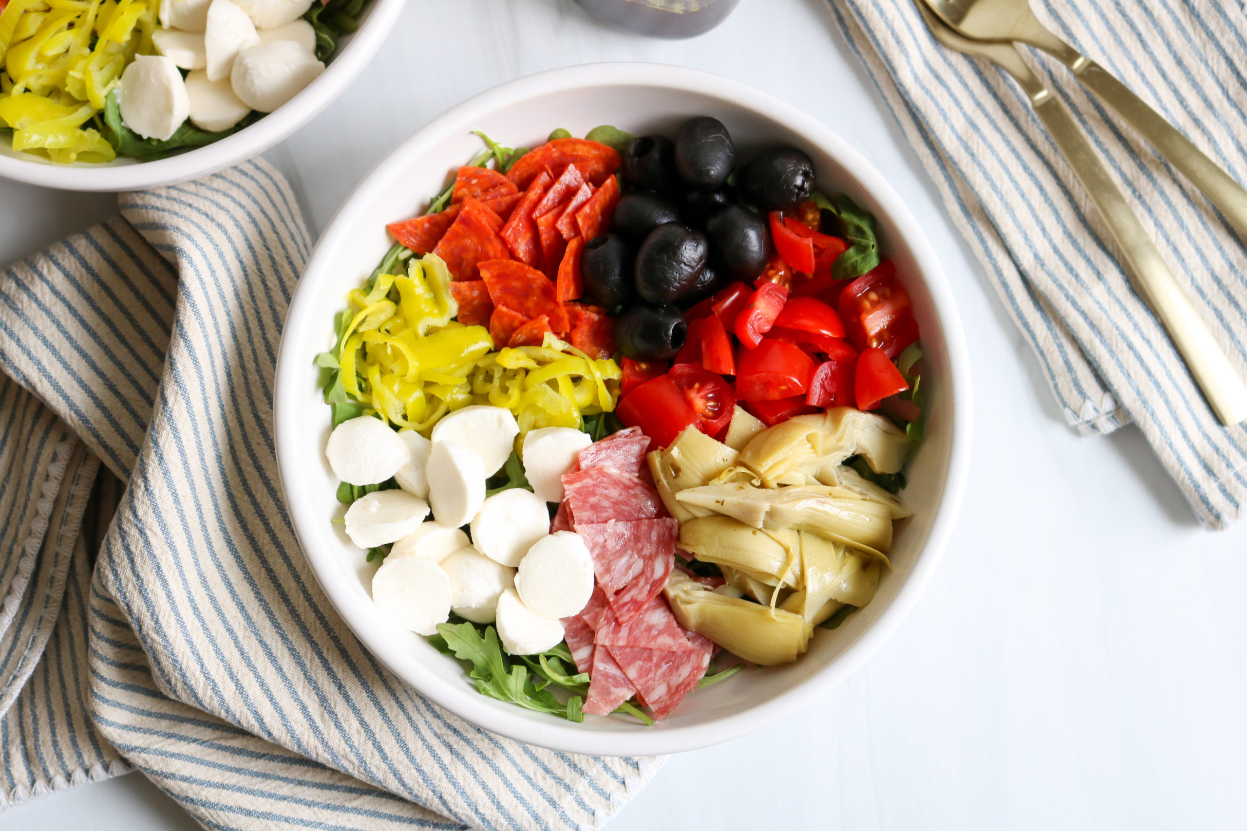 A large white serving bowl filled with antipasto salad including chopped romaine, salami ribbons, provolone cubes, cherry tomatoes, pepperoncini, olives, and red onion on a bright kitchen counter