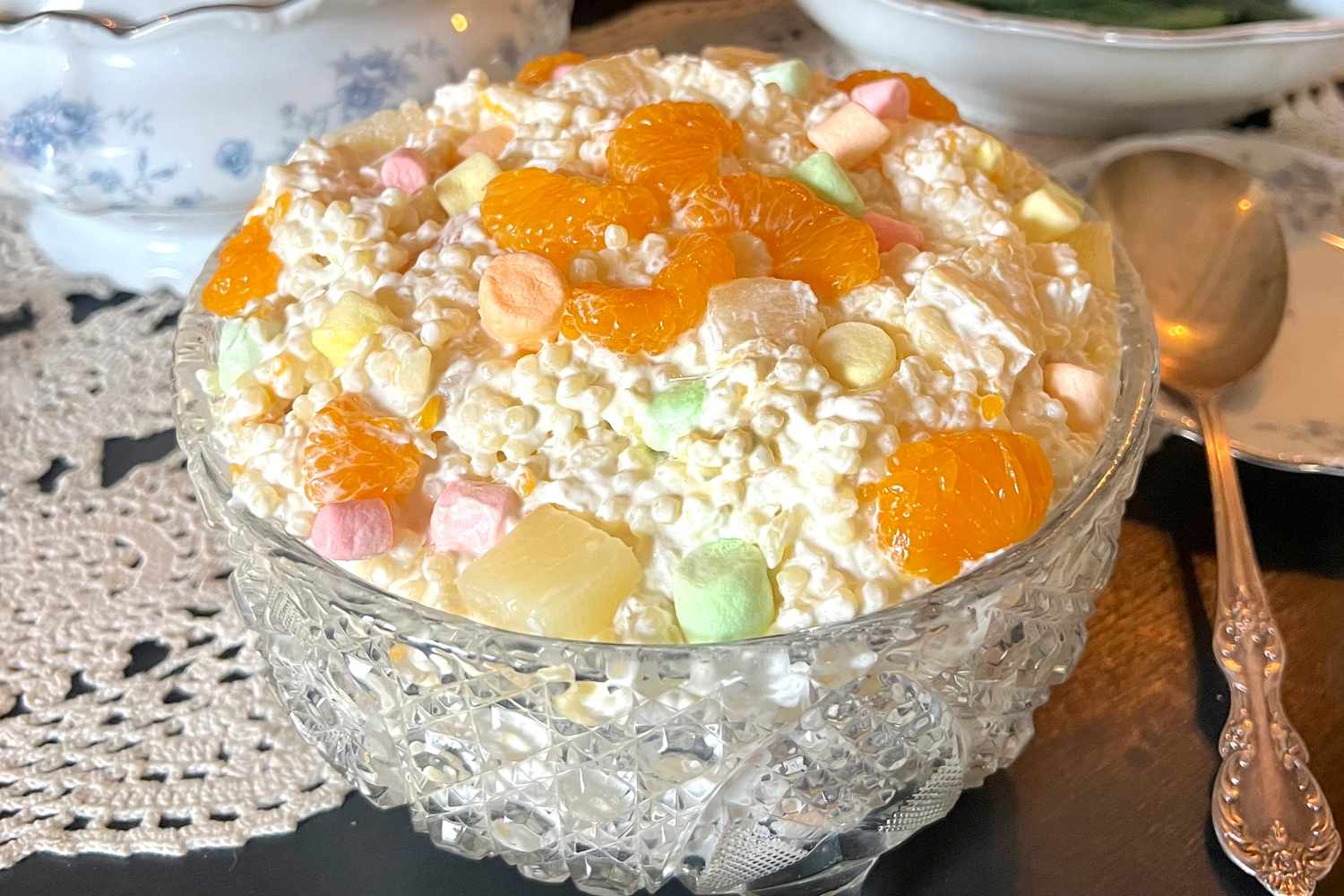 A large white serving bowl filled with creamy frog eye salad with visible acini di pepe pasta, mandarin orange segments, pineapple pieces, mini marshmallows, and a sprinkle of shredded coconut on a bright kitchen table