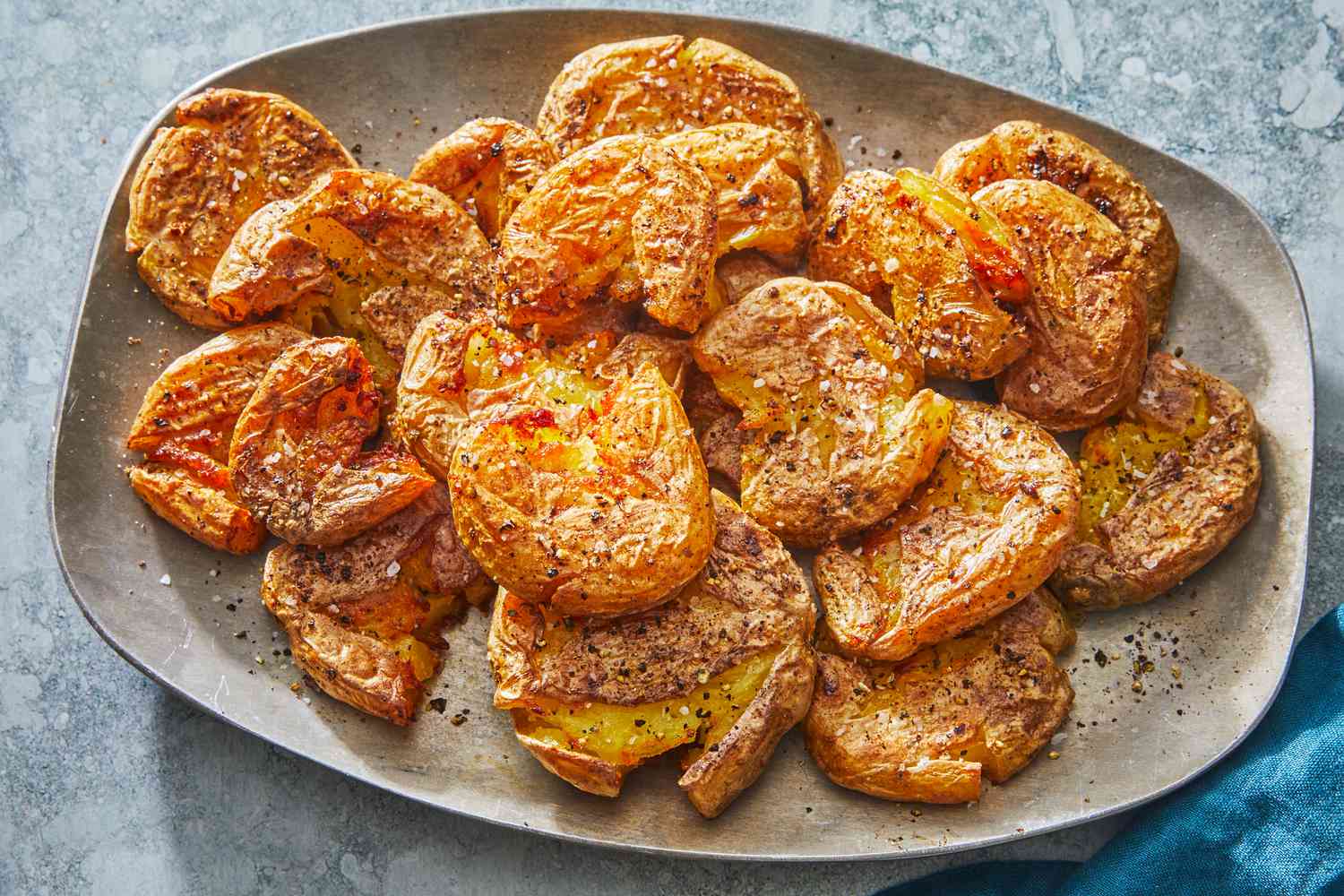A large white serving platter piled with golden crispy smashed baby potatoes, sprinkled with chopped herbs and flaky salt on a bright kitchen table