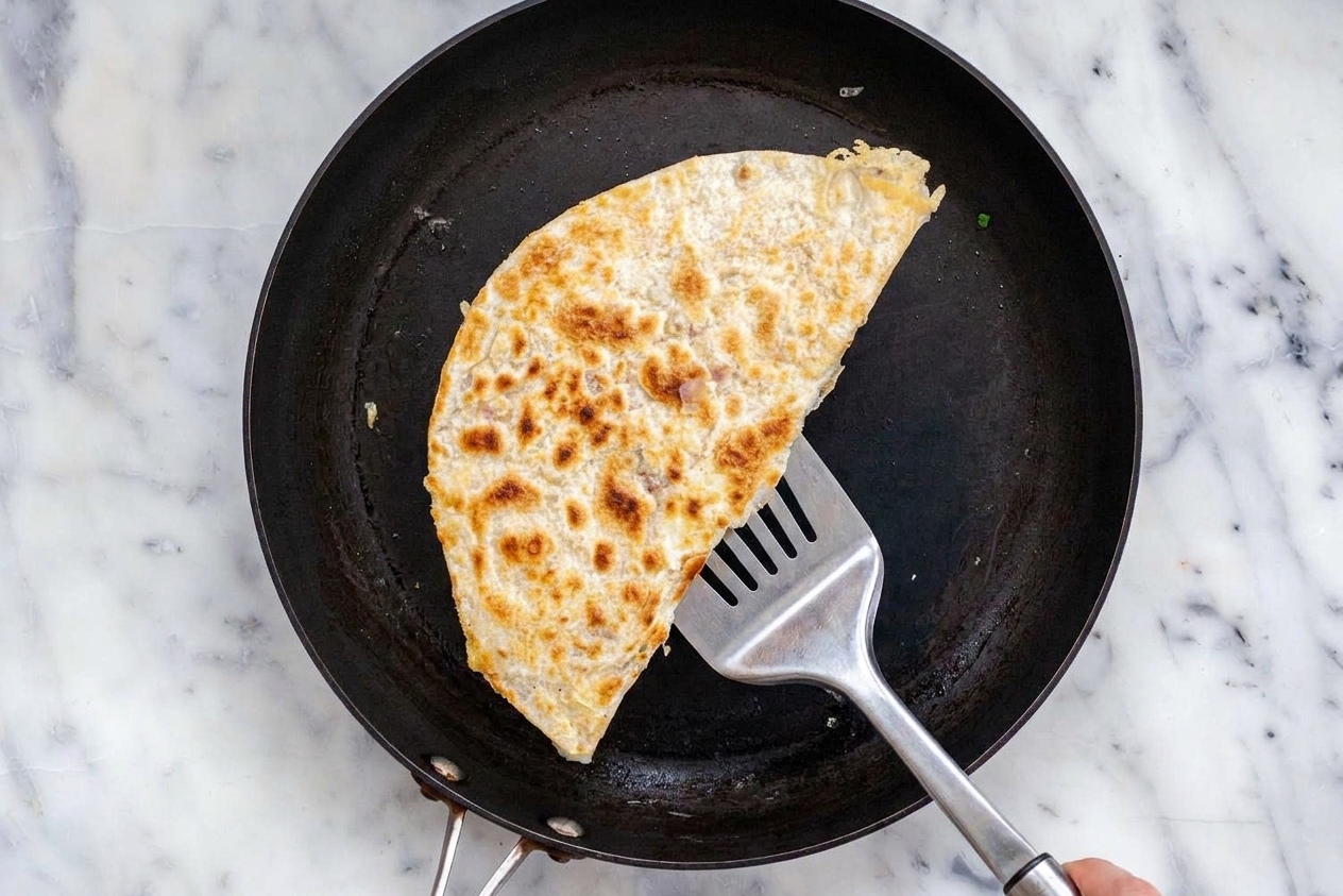A leftover quesadilla wedge reheating in a nonstick skillet with a spatula lifting one corner to check browning