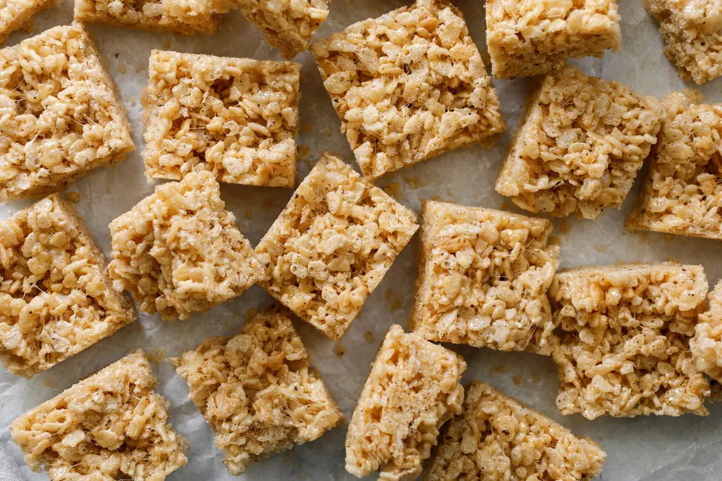 A lightly buttered knife slicing warm peanut butter Rice Krispie treats in a parchment-lined square pan on a wooden counter
