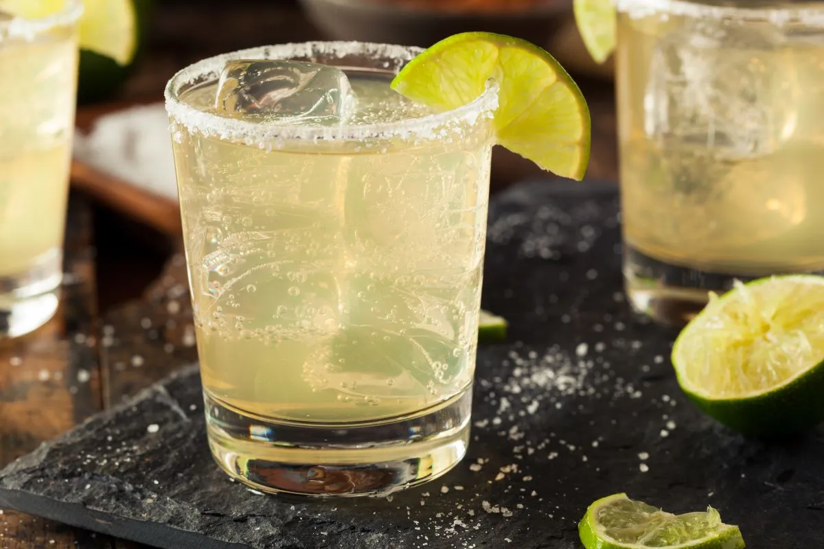 A lime wedge and a small plate of kosher salt next to a rocks glass being rimmed on a kitchen counter