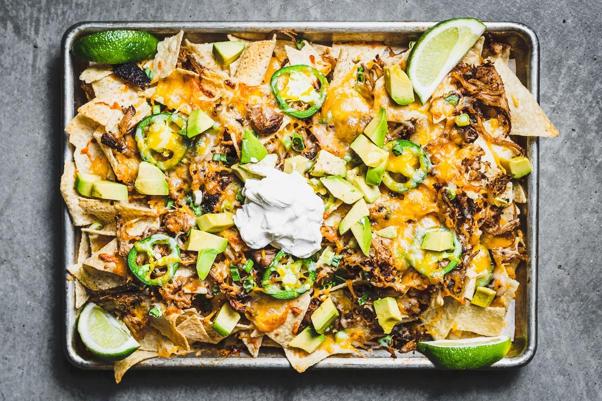 A loaded sheet pan of pulled pork nachos with melted cheddar and Monterey Jack, caramelized pulled pork, jalapeño slices, cilantro, and dollops of sour cream on a parchment-lined pan in a warm home kitchen