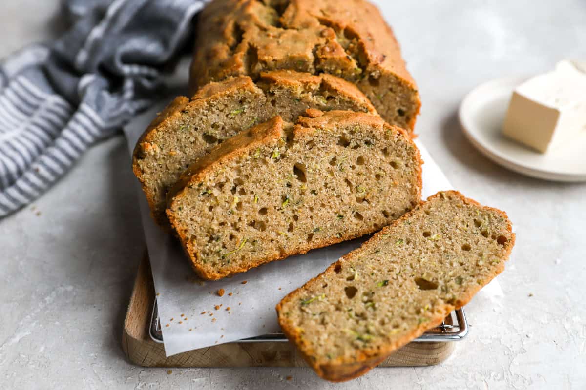A loaf of zucchini bread cooling on a wire rack with a few slices cut and crumbs on a cutting board