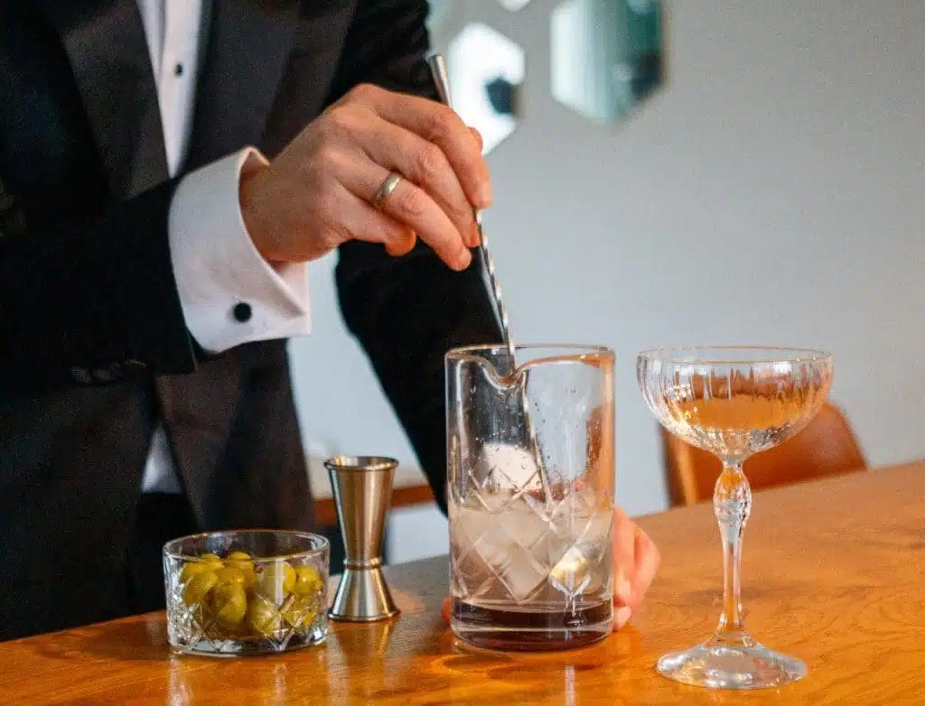 A long bar spoon stirring gin and vermouth in a clear mixing glass packed with ice on a stone countertop under soft kitchen lighting