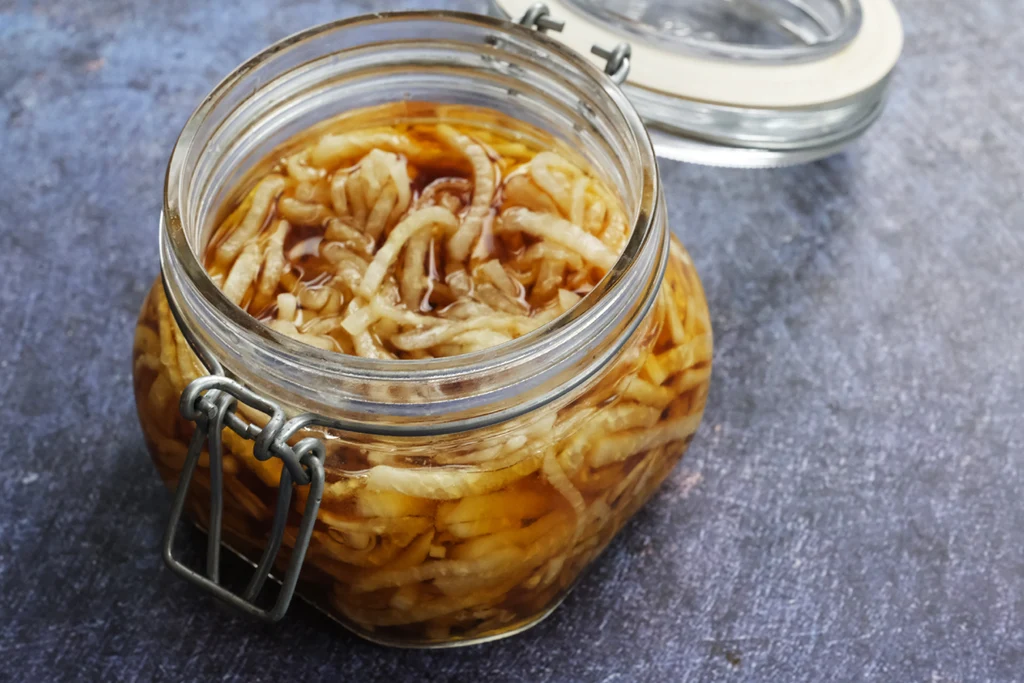 A mason jar filled with thin matchsticks of quick pickled daikon in clear brine, sitting on a light kitchen counter with soft natural window light