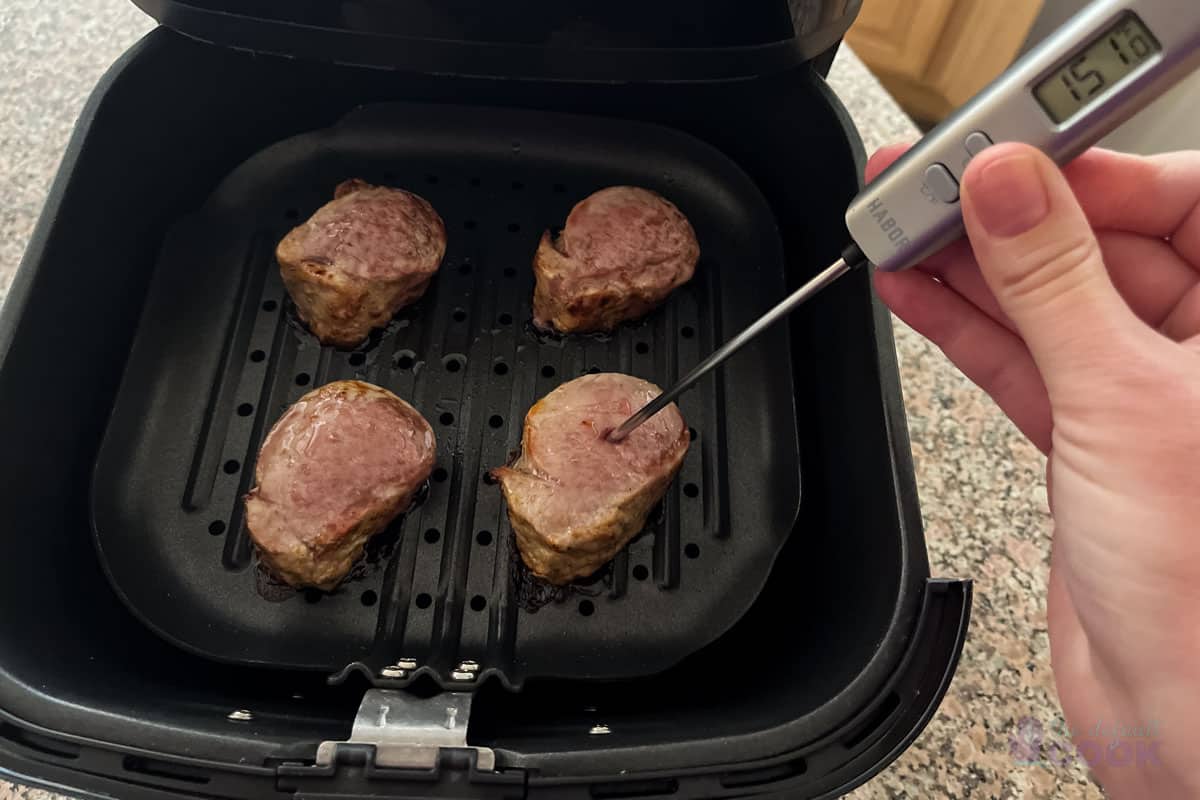 A meat thermometer inserted into the thickest part of a cooked pork tenderloin in an air fryer basket with a browned spice crust