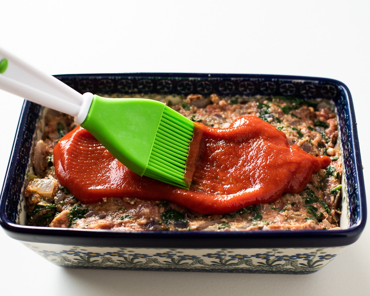 A meatloaf on a parchment-lined sheet pan being brushed with a thick ketchup glaze using a silicone brush, warm kitchen lighting