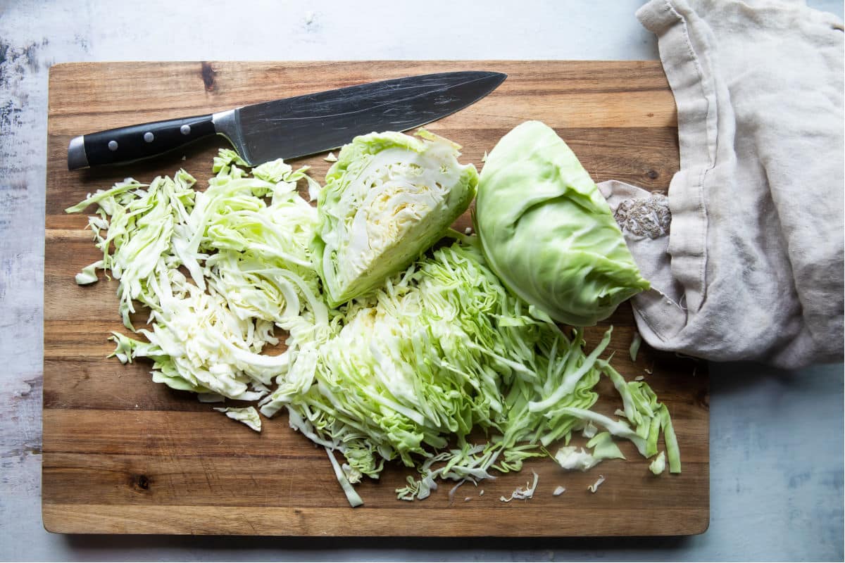 A medium green cabbage cut into wedges on a wooden cutting board with a chef’s knife nearby, in a bright home kitchen