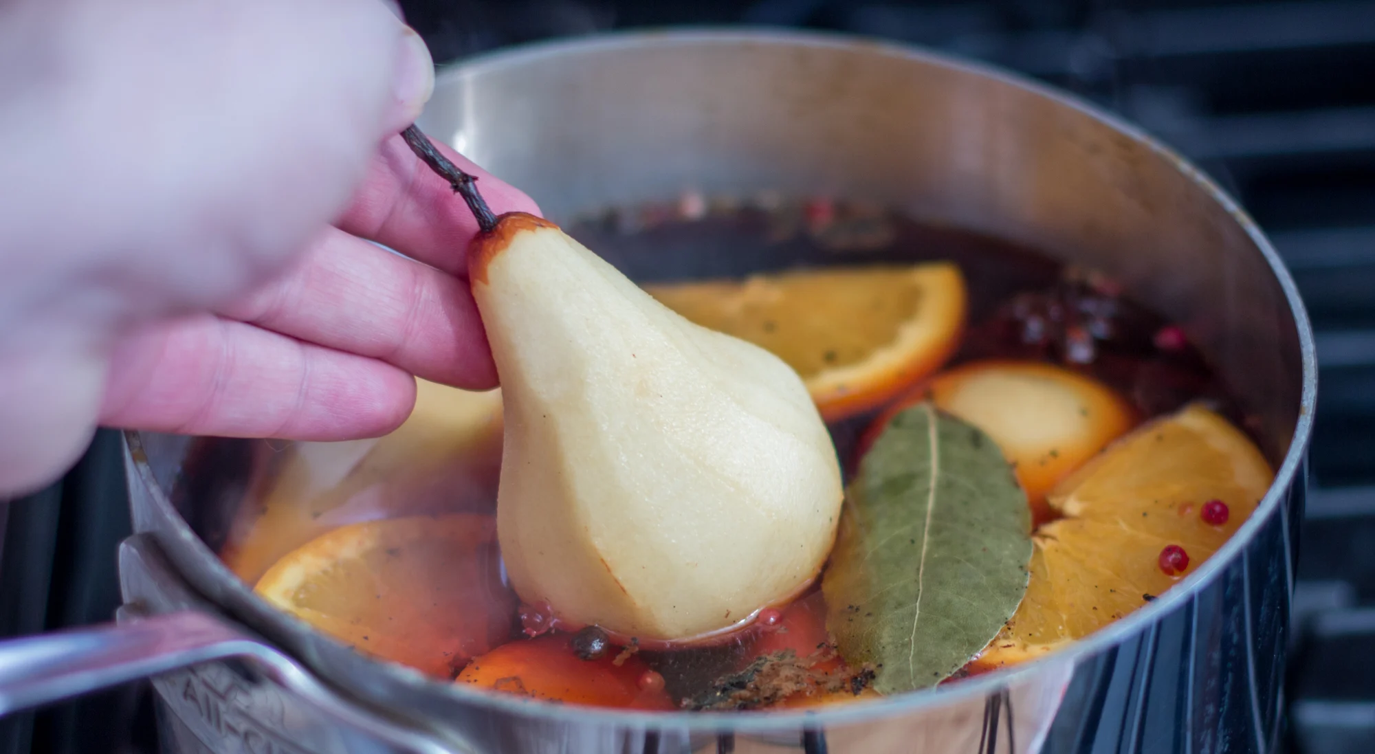A medium pot on a stovetop with pears gently simmering in red wine, cinnamon sticks floating on the surface, and light steam rising in a cozy kitchen
